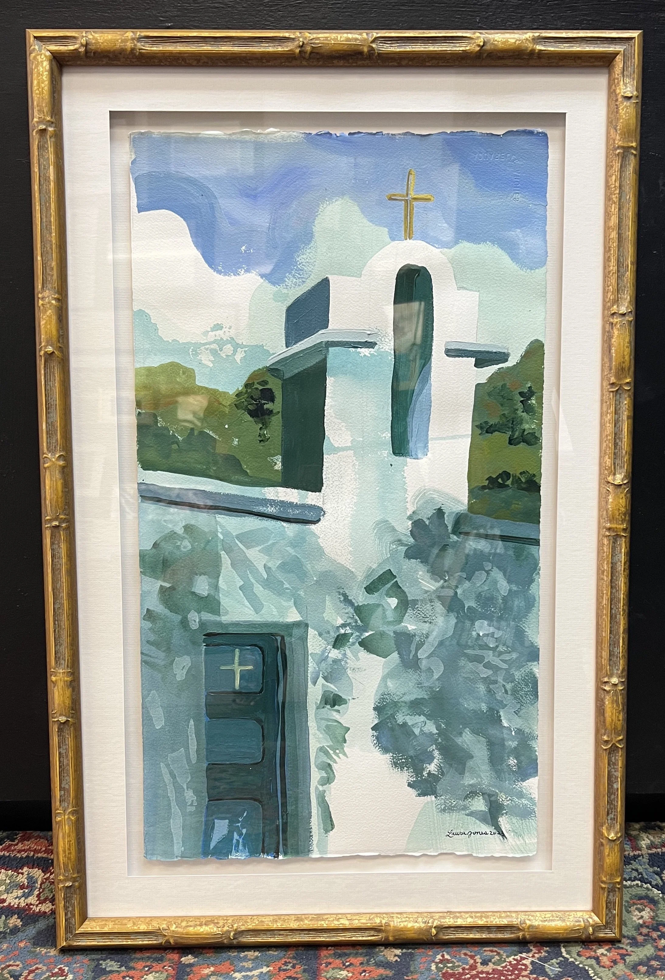 Framed painting of a church with a prominent tower and cross at the top, surrounded by greenery, using shades of blue and green, in a bamboo-style frame.