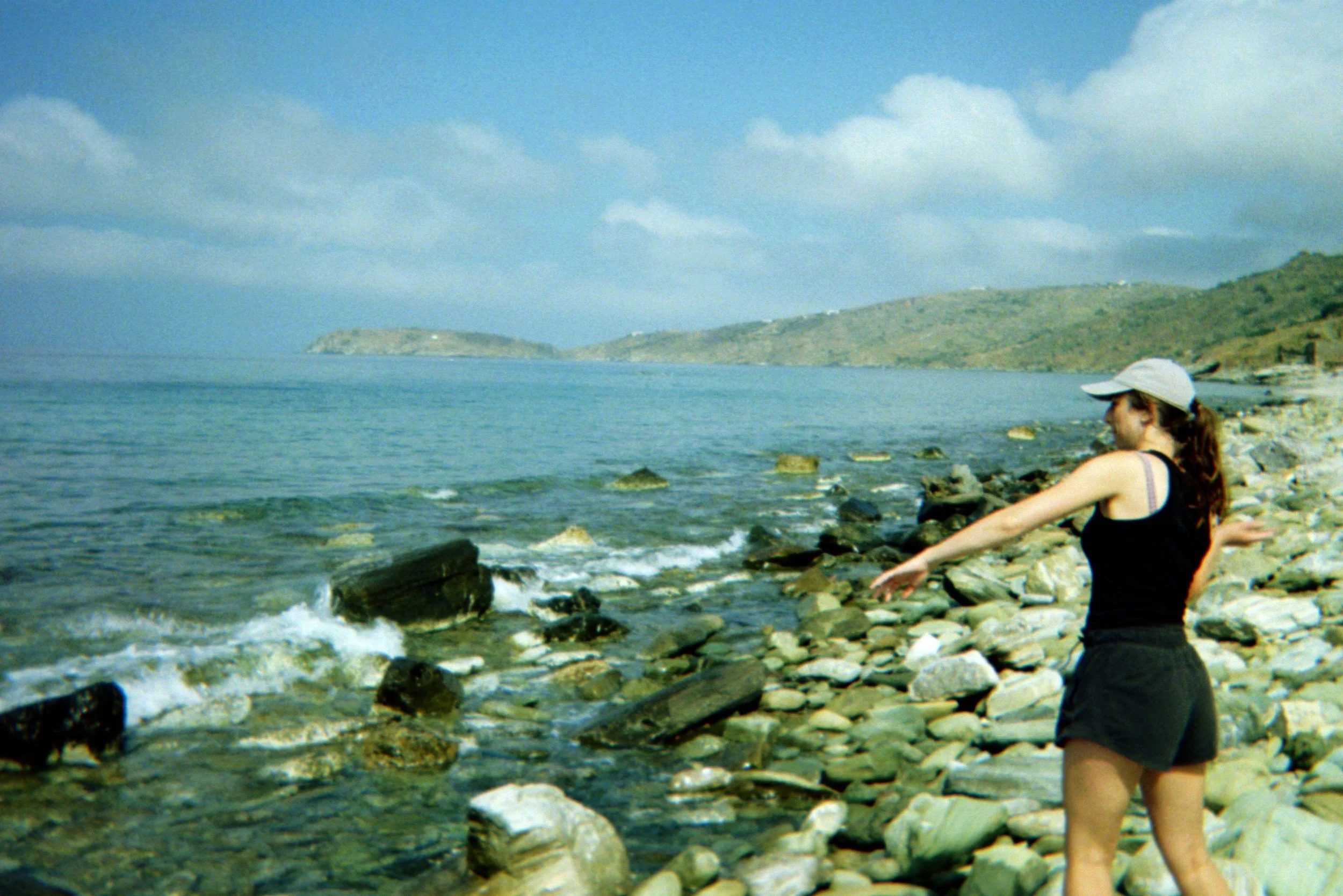 Person skipping rocks on a rocky beach with a view of the ocean and distant hills under a partly cloudy sky.