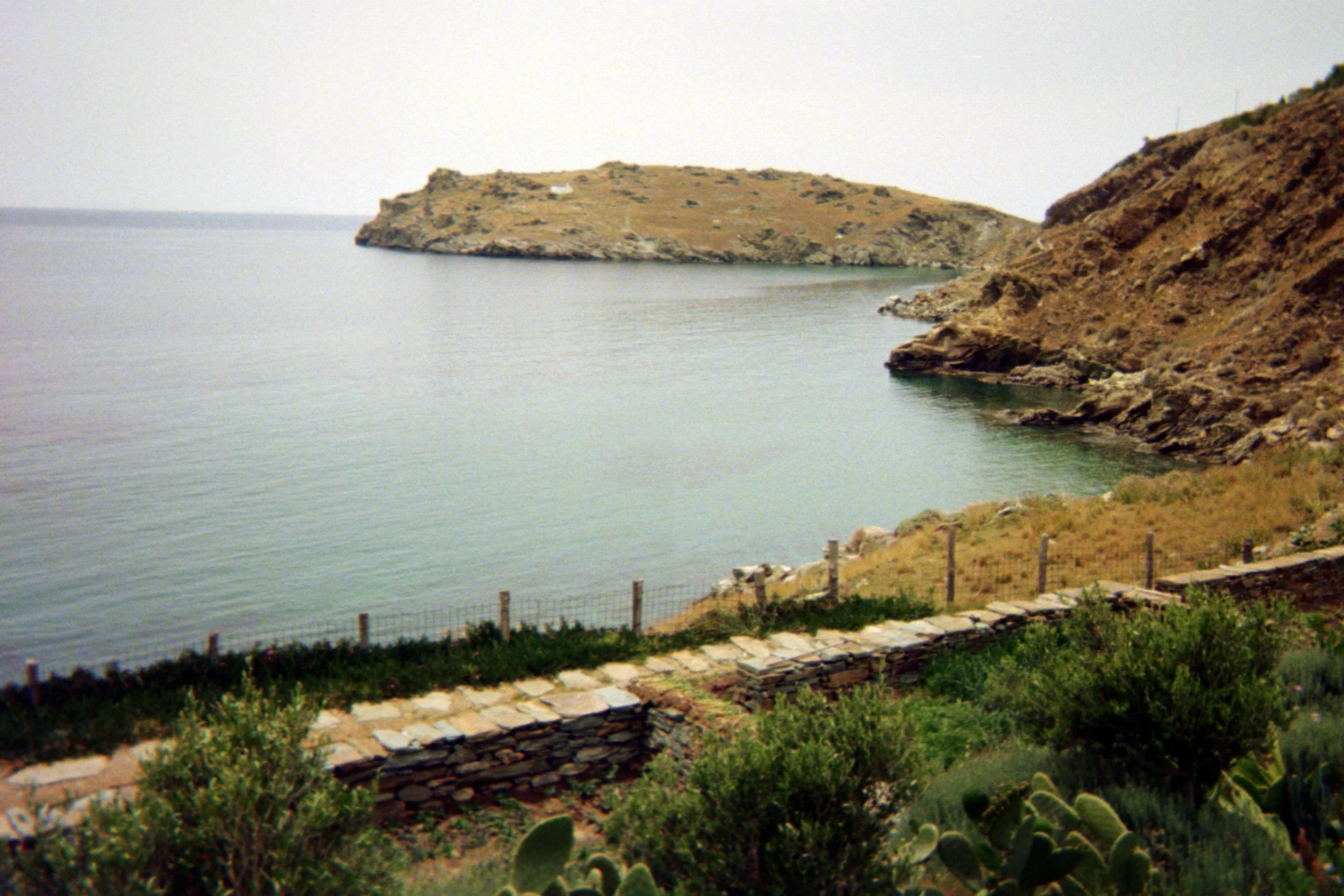 Rocky coastal landscape with calm sea and a stone pathway, surrounded by sparse vegetation, leading to a small fenced garden area.