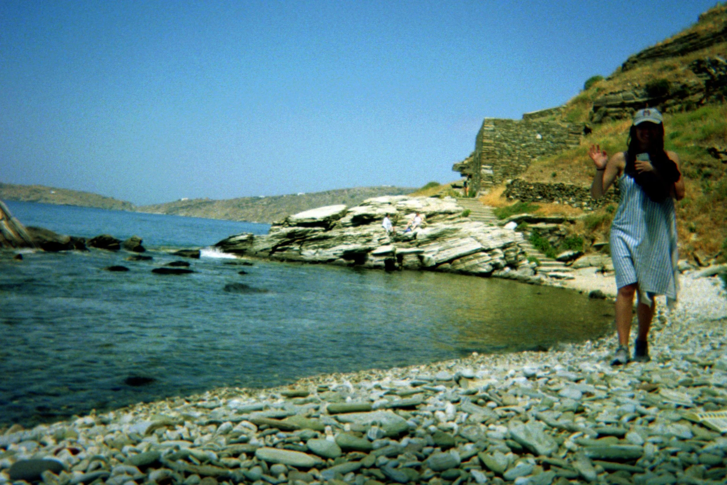 Rocky beach with a woman in a striped dress and hat walking along the shore, a stone structure in the background, and clear blue water.