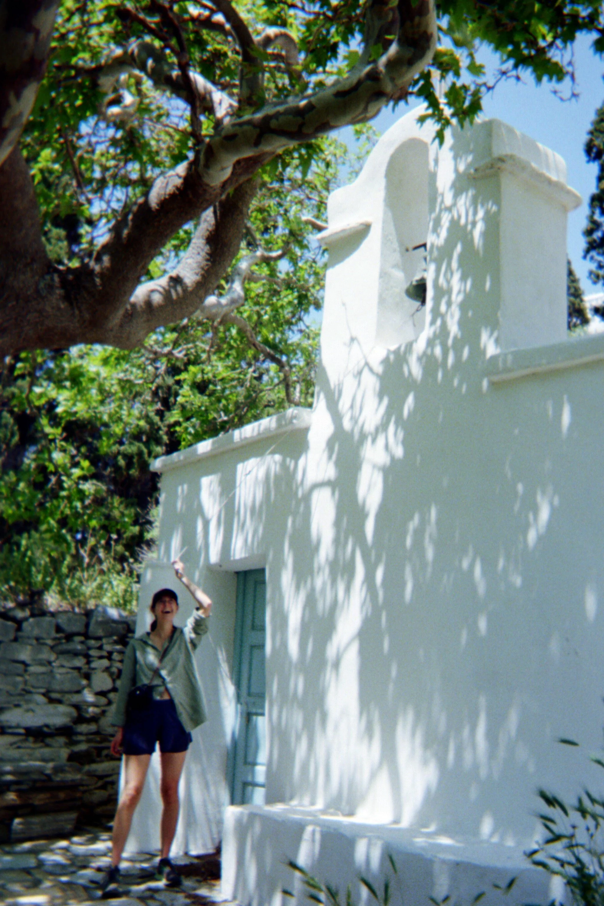Person standing outside a white building with a small bell tower, surrounded by lush green trees and shadows.