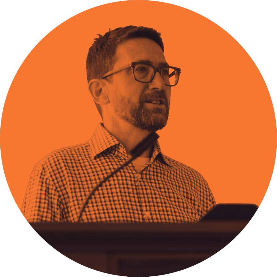 Man with glasses and a checkered shirt speaking at a podium with an orange background.