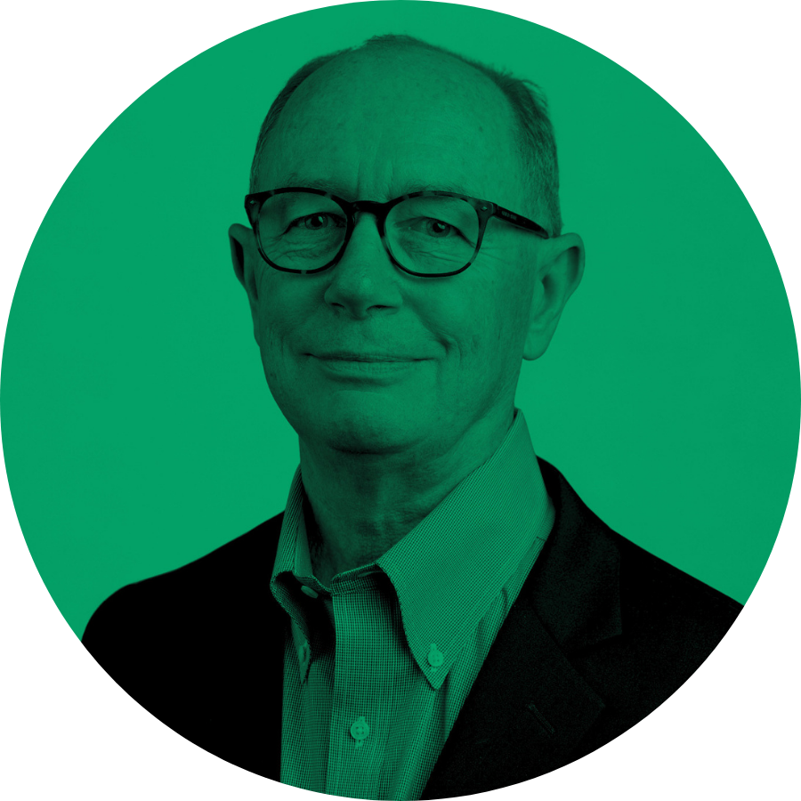 A middle-aged man with glasses, wearing a green collared shirt and a black blazer, is looking at the camera with a slight smile.