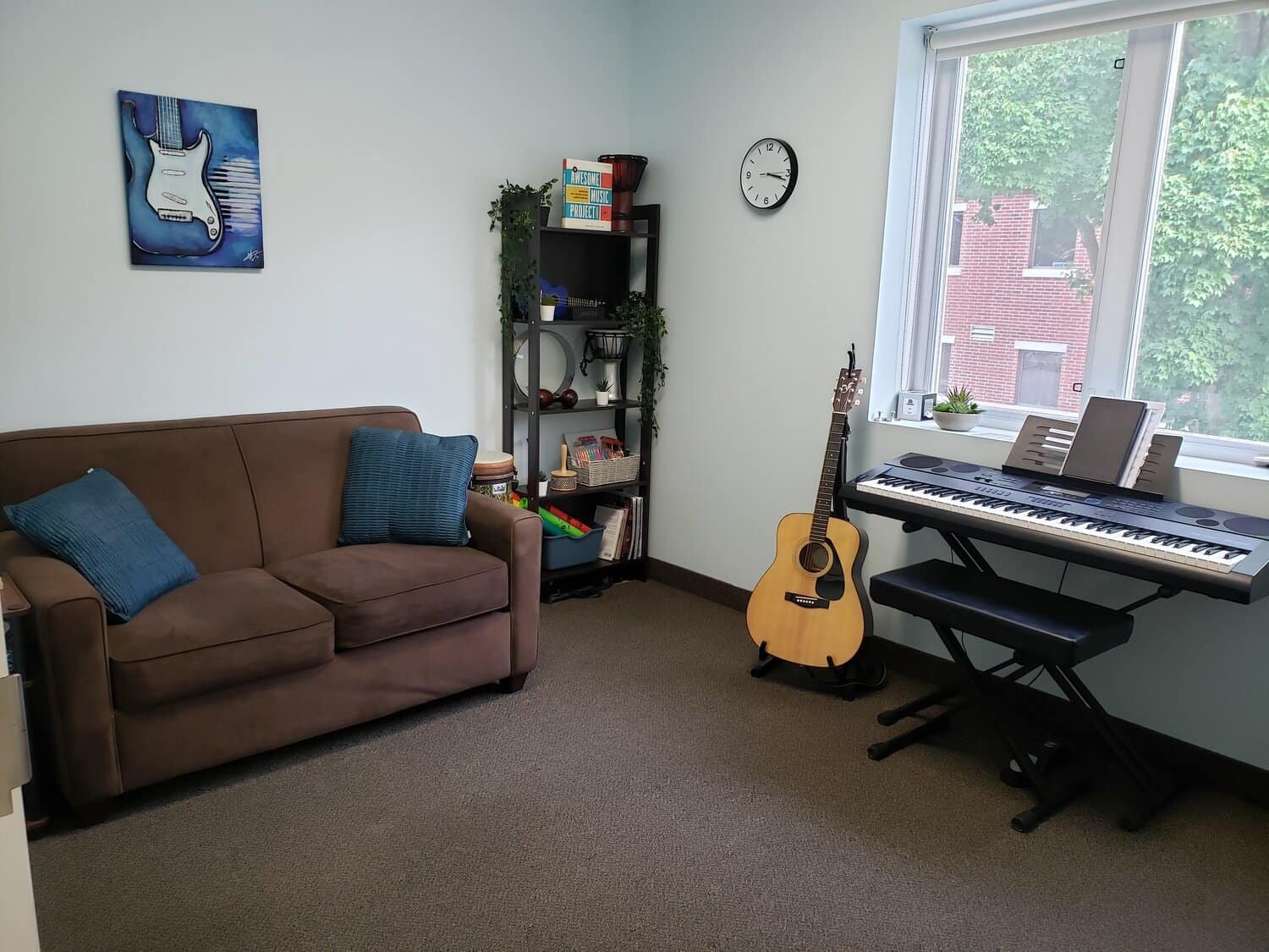 A cozy music room with a brown sofa, blue cushions, and a painting of a blue electric guitar on the wall. A clock is mounted above a bookshelf with books, a guitar, and various items. There's a window with a view of trees, a digital keyboard on a sta