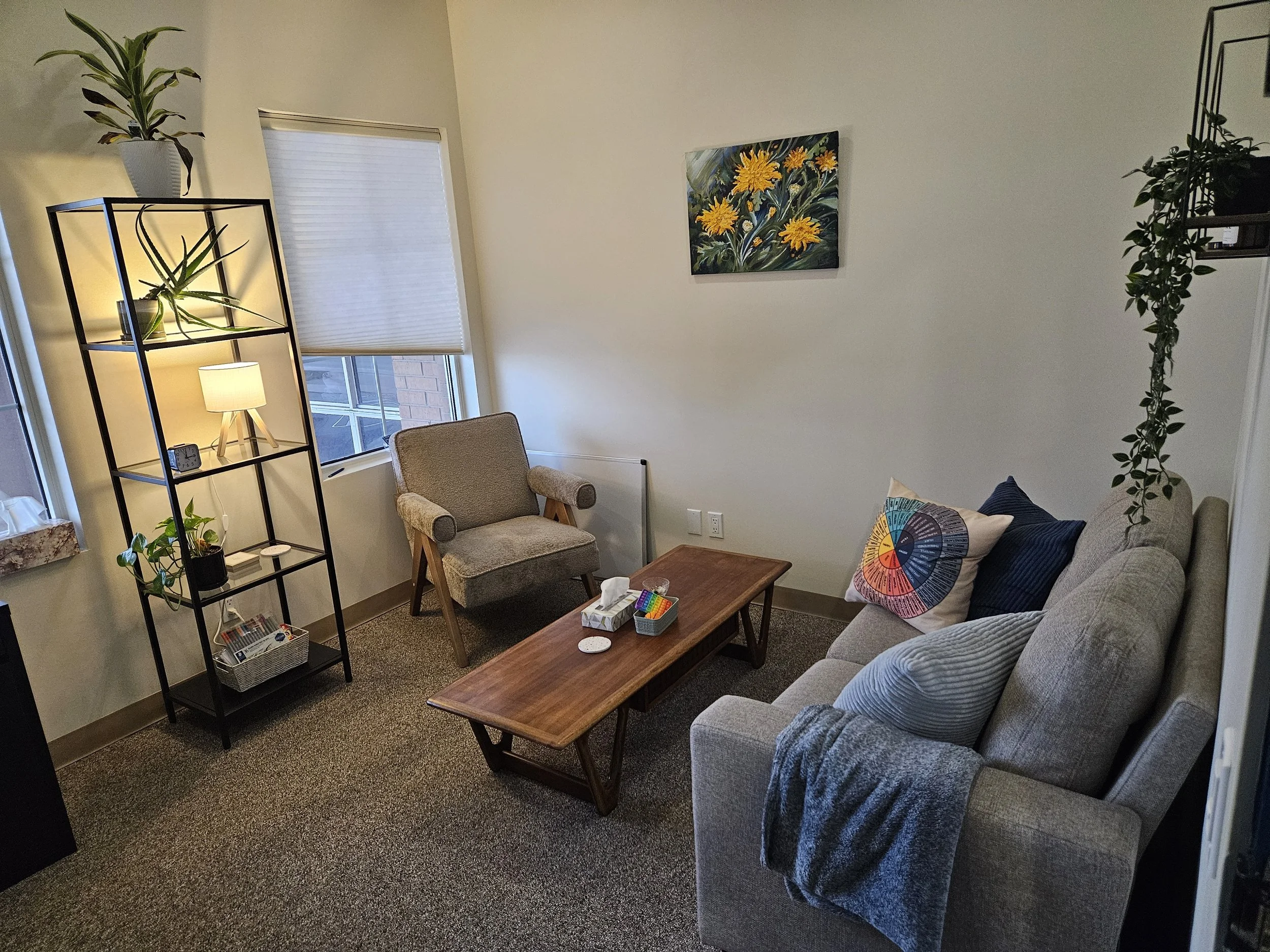 A cozy counselling office with a beige sofa, a gray armchair, and a wooden coffee table. There are decorative pillows on the sofa, a colorful circular wall art, and some hanging plants. 