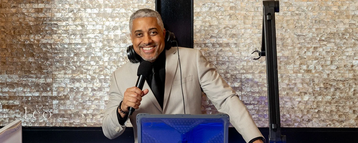 a smiling man in a suit hosting a radio or podcast show with headphones, microphone, and equipment in a studio setting