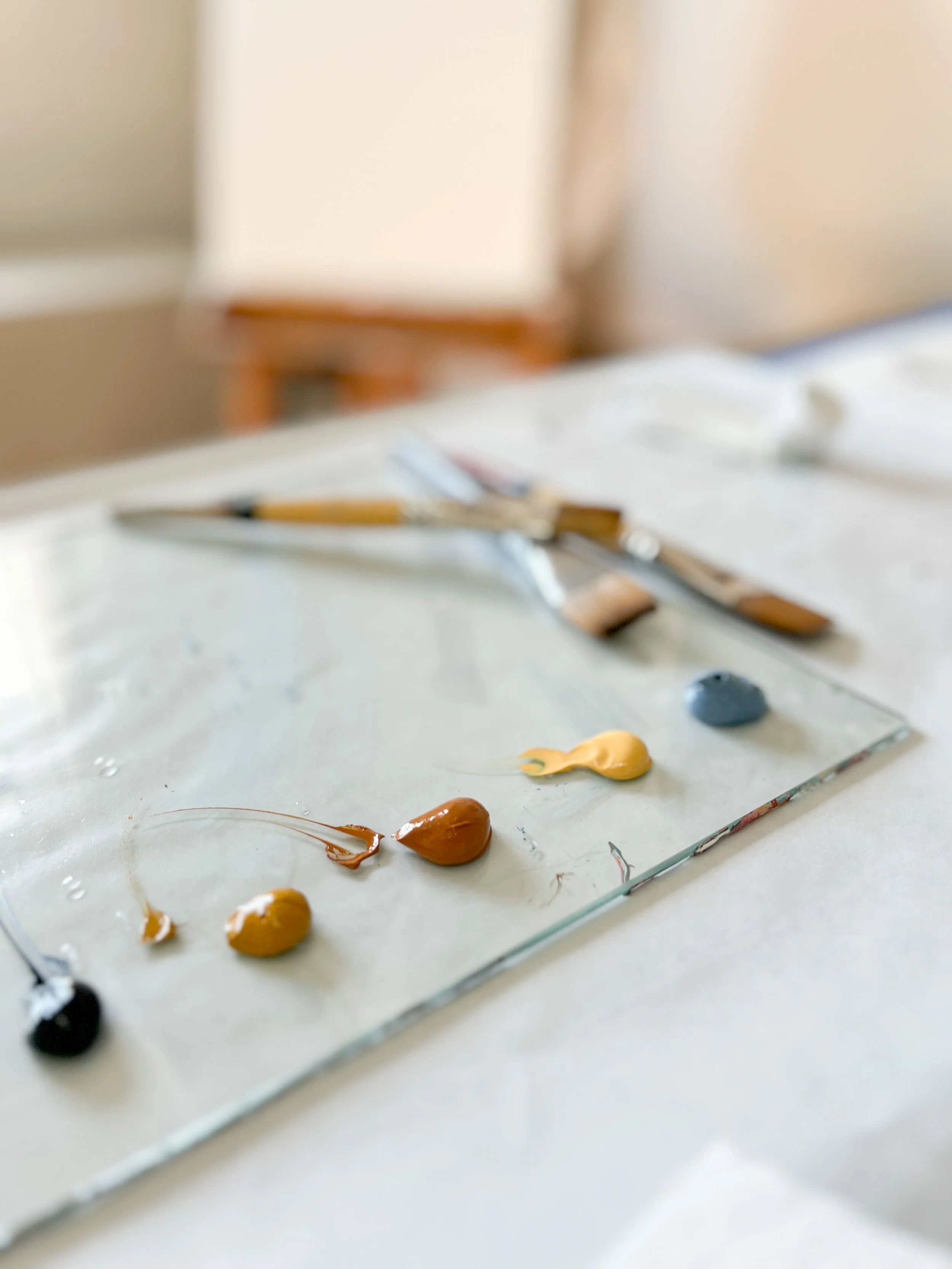 Paint palette with dollops of various paint colors, including brown, yellow, blue, and black, on a glass surface. Paintbrushes are placed in the background.