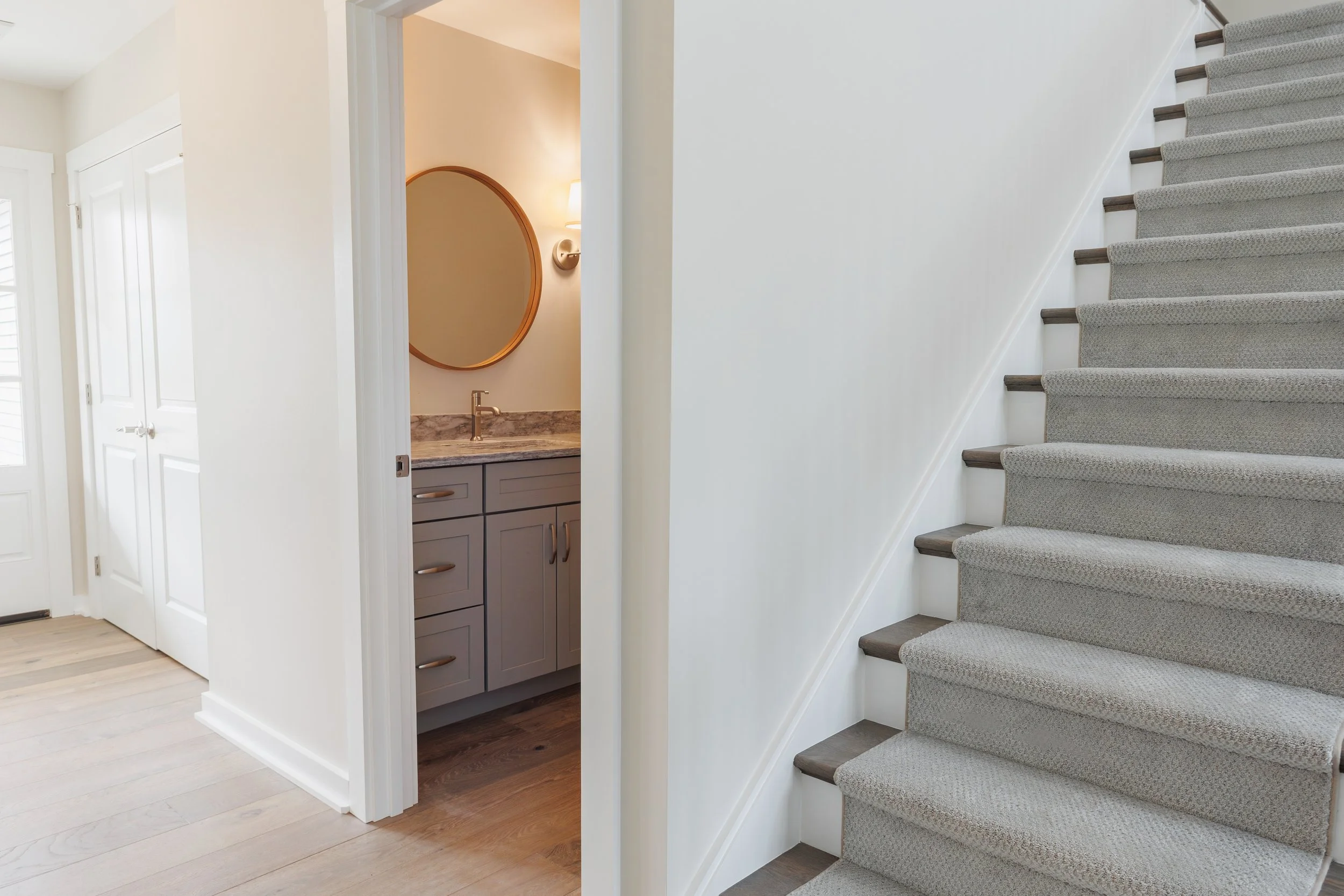Modern house interior with a staircase, a small bathroom with a round mirror and vanity, and white walls.