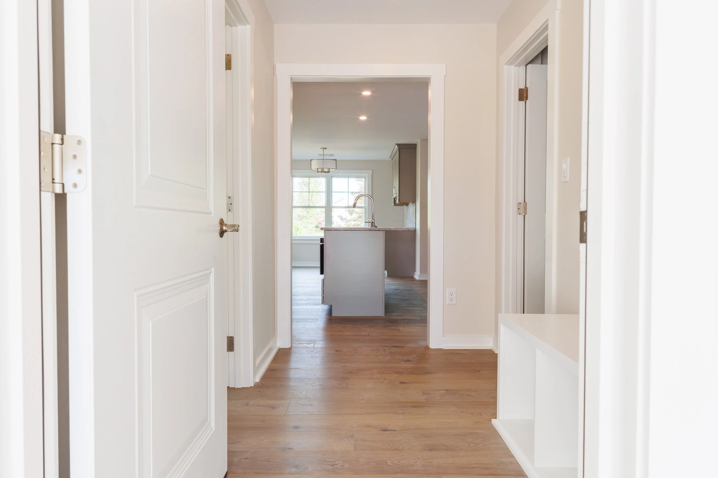 Interior view of a hallway leading to a kitchen with a window, wooden floors, white walls, and ceiling with recessed lighting.