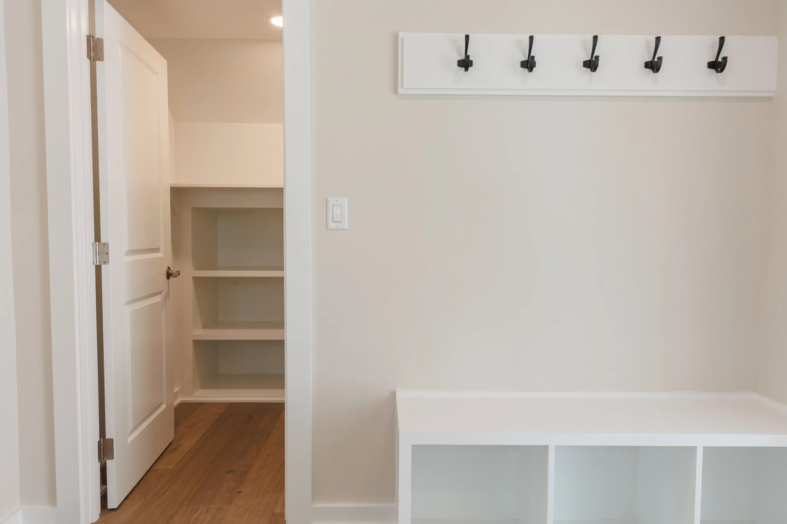 Empty built-in white shelving unit with open cubbies and a row of black hooks on the wall, a door open to a small closet or storage space, and wood flooring in a room with light-colored walls.