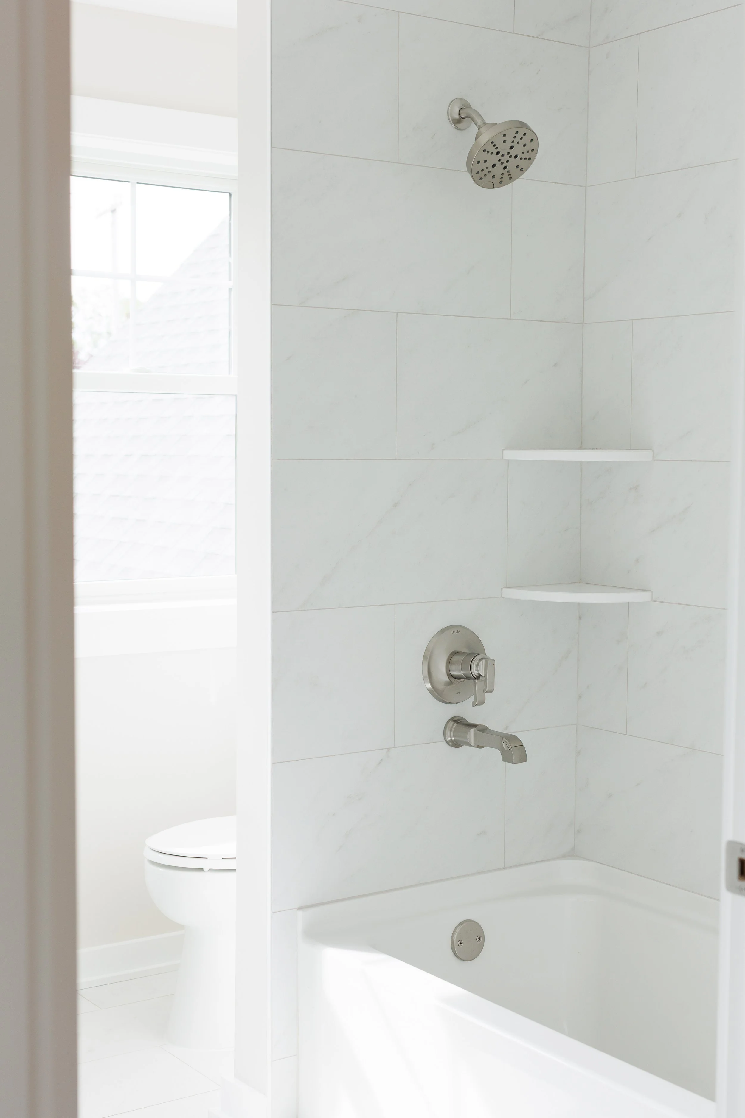 A bathroom shower with white tiled walls, a wall-mounted showerhead, a metal faucet, and a small built-in corner shelf. A window with a view of the outside is partially visible.