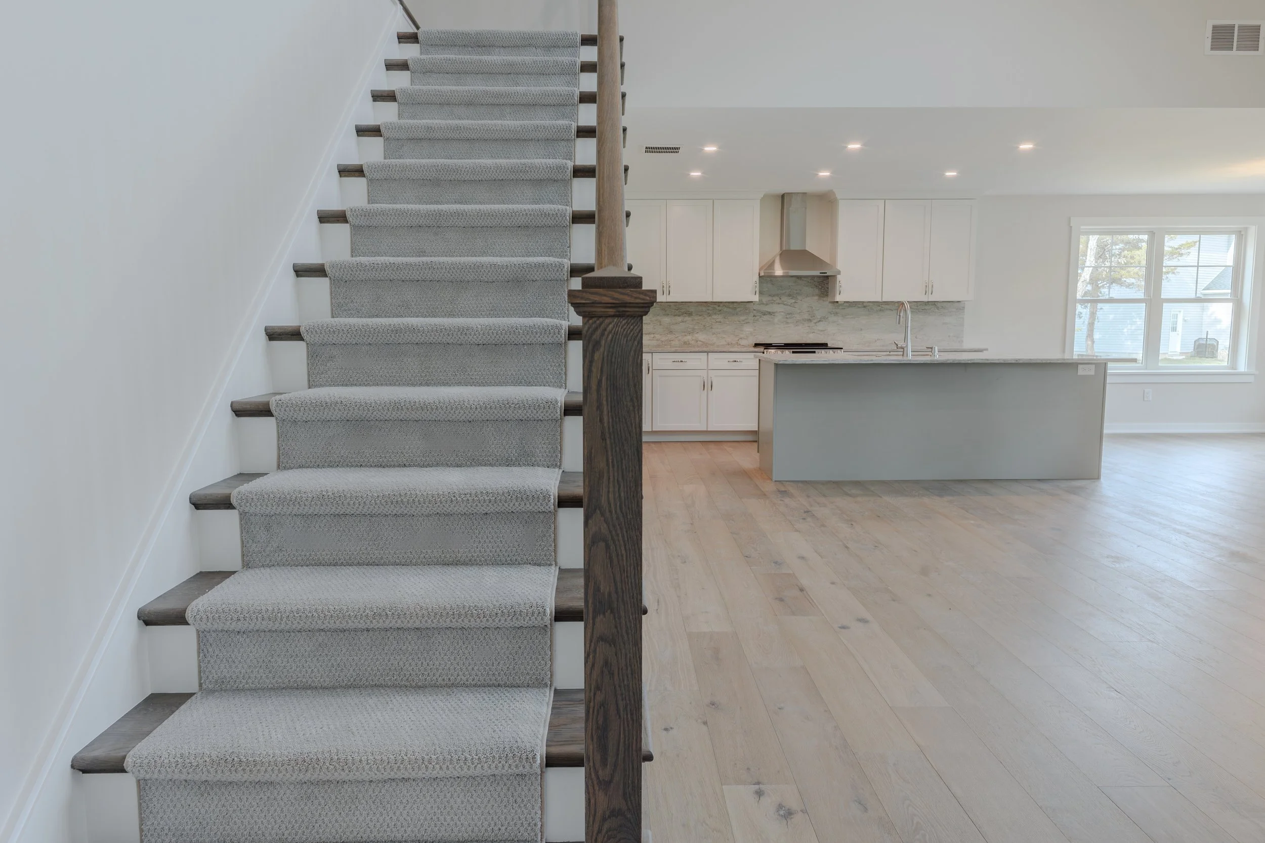 Interior view of a modern open-concept kitchen and living space with a staircase on the left. The staircase has carpeted steps, wooden treads, and a wooden handrail. The kitchen has white cabinets, a marble backsplash, and a large window letting in n