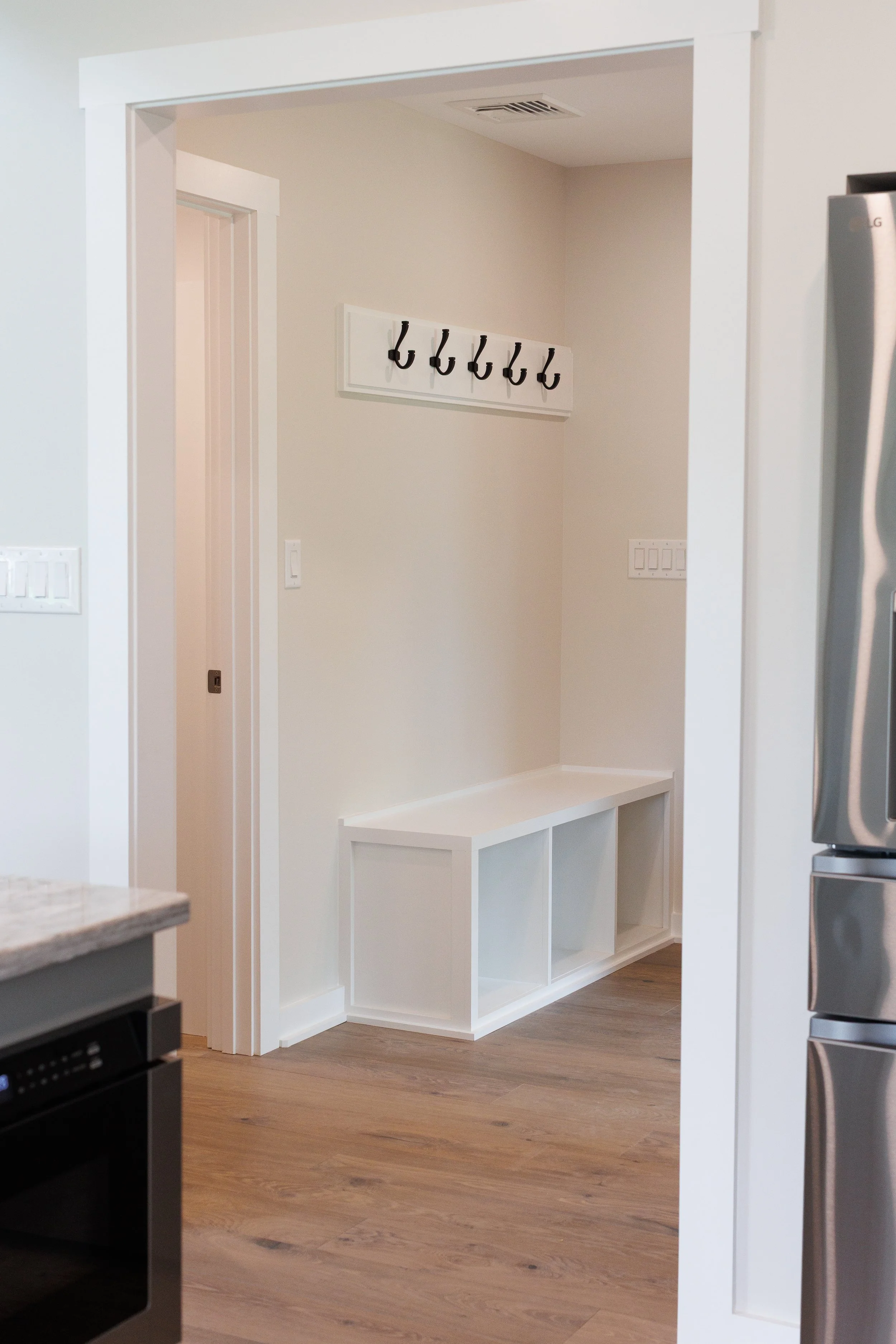 A wall area in a home with a white built-in coat rack and cubby storage beneath it, adjacent to a doorway, with a beige wall and wooden floor.