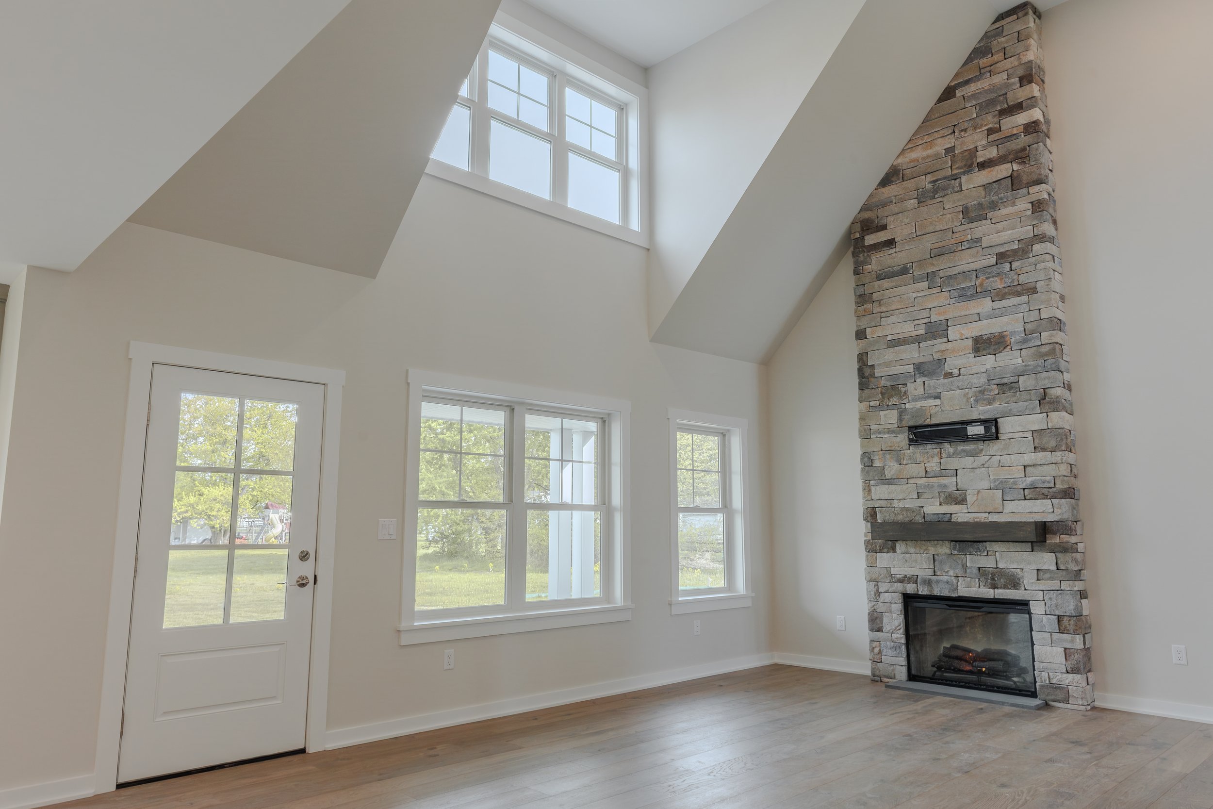 A bright living room with large windows, a stone fireplace, white walls, and hardwood floors, with a door leading outside.