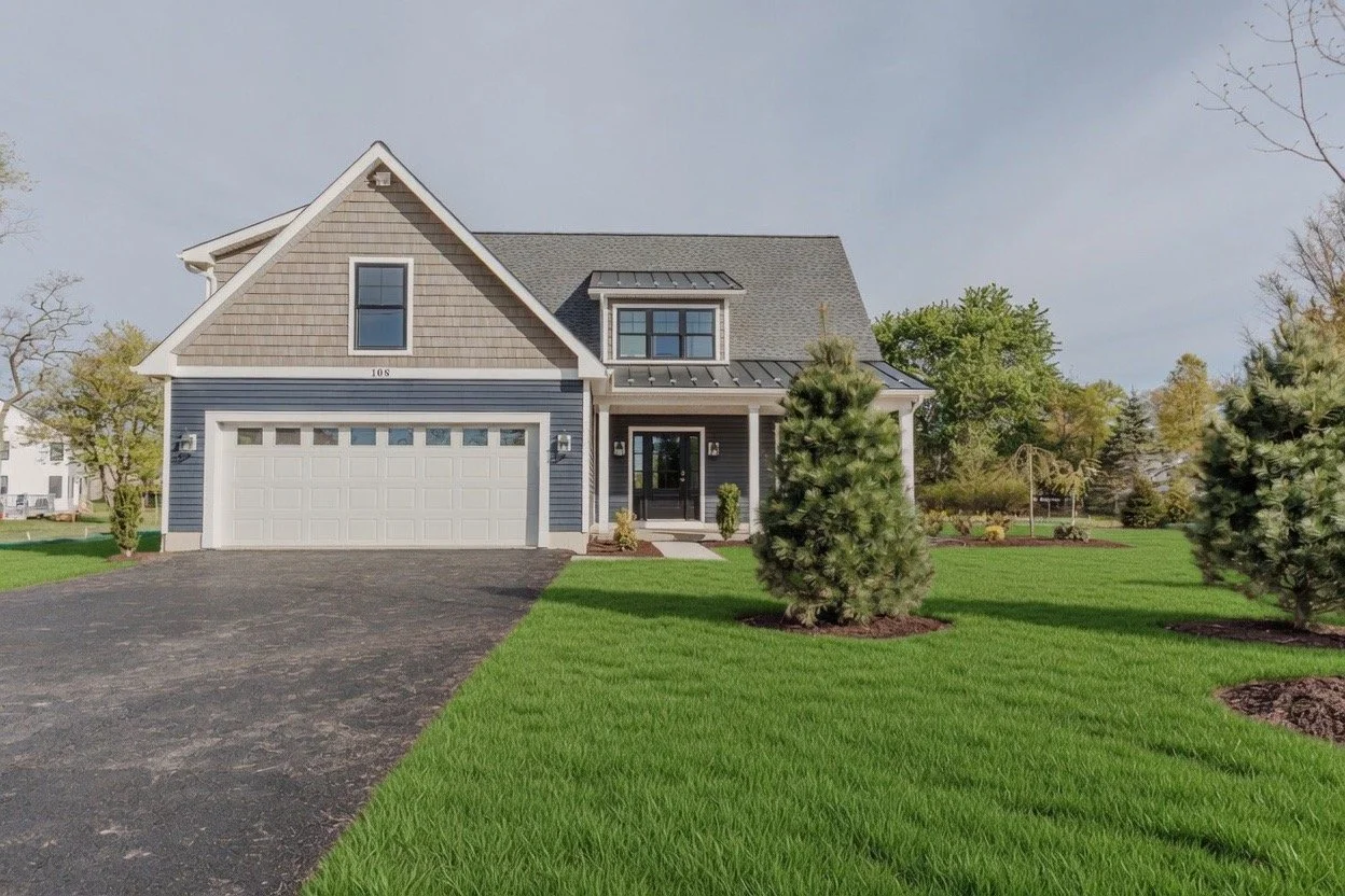 Front view of a modern two-story house with a garage, blue and beige siding, front porch, landscaped yard with small trees and green grass.