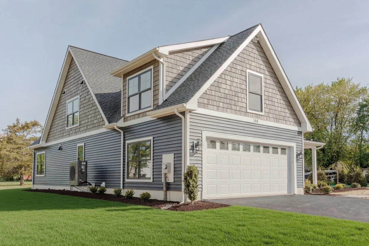 A two-story house with gray siding and shingle accents, white trim, a garage door, and a small front lawn with shrubs.