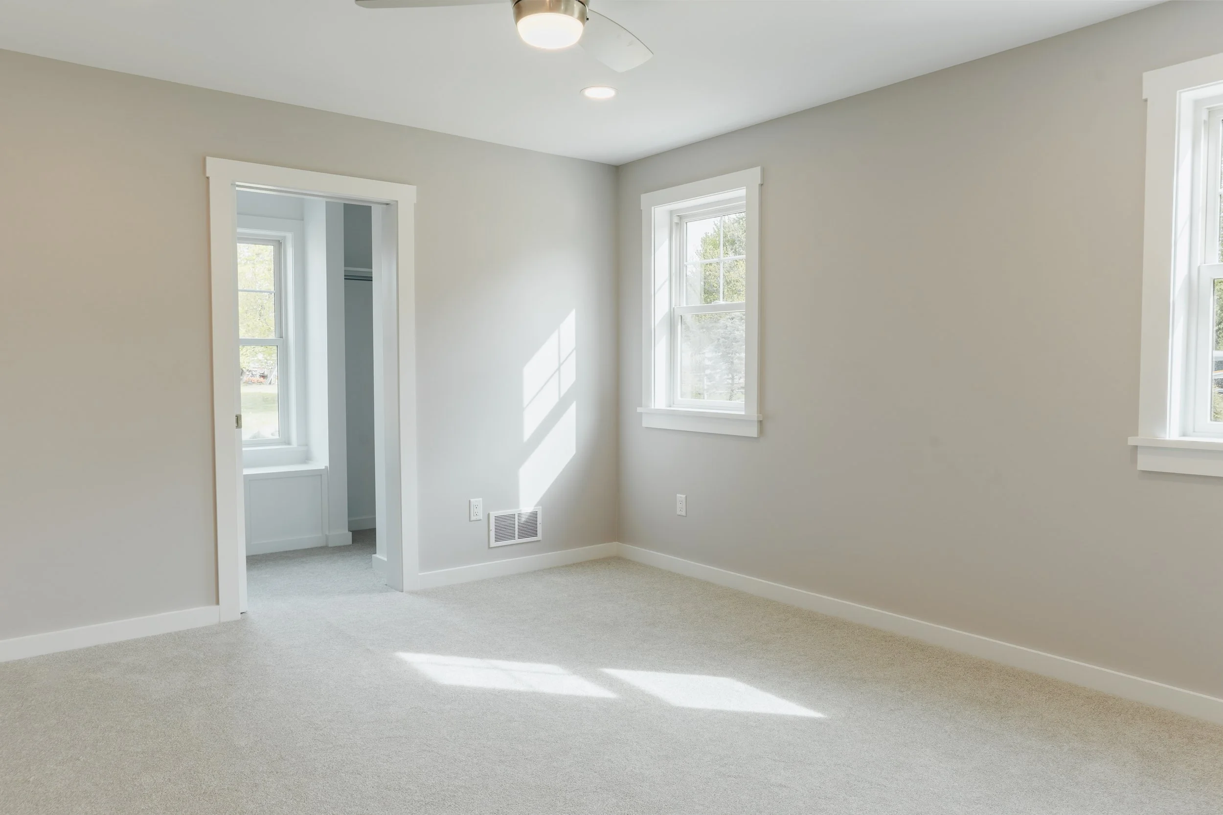 Empty room with white walls, three windows allowing natural light, and beige carpet floor. Ceiling with a mounted light fixture and a ceiling fan.