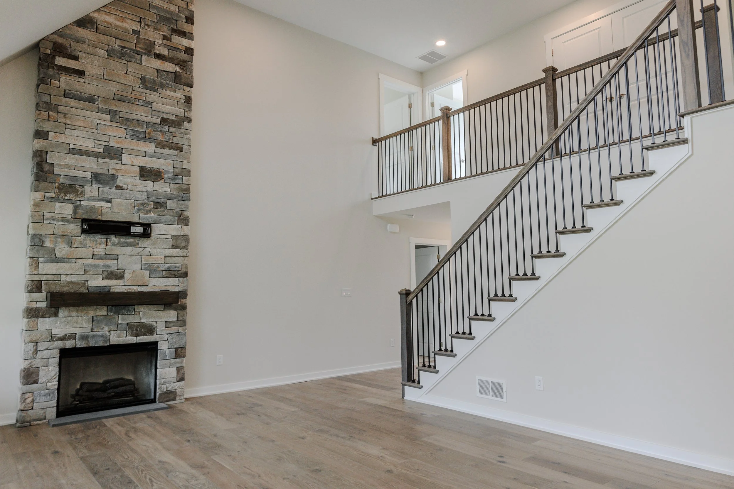 Interior of a modern home with a stone fireplace, hardwood floors, and a staircase with black metal and wooden handrails leading to an upper level.
