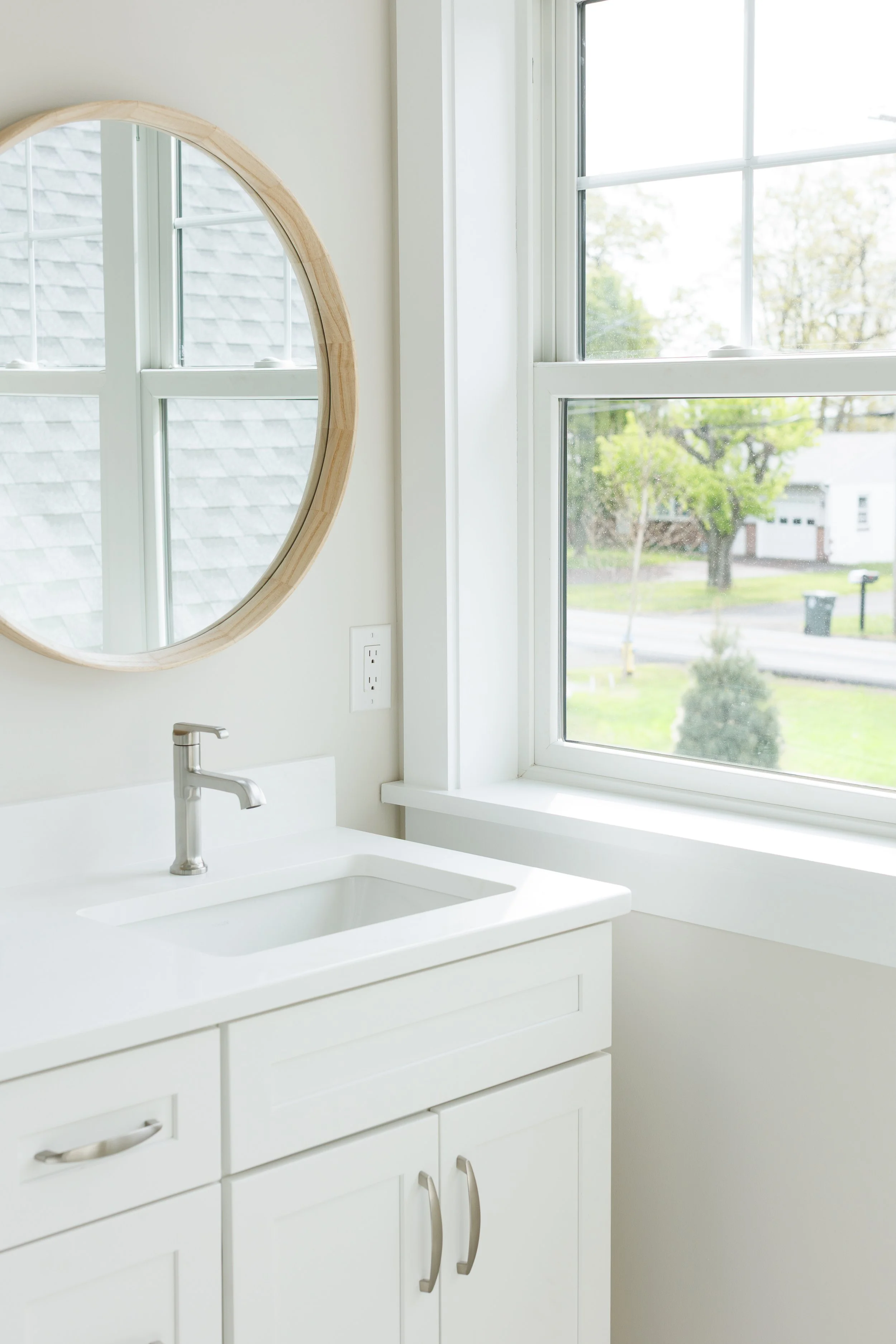 A white bathroom vanity with a stainless steel faucet, a round wooden-framed mirror, and a large window showing trees and houses outside.