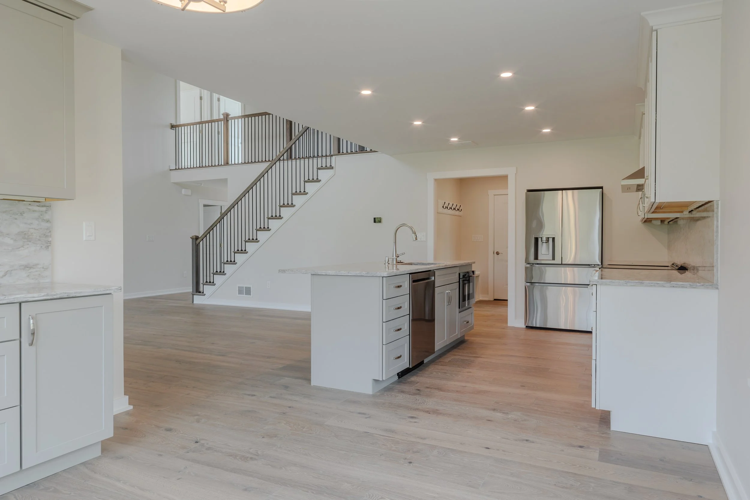 Open-concept kitchen and living room with stainless steel refrigerator, island, staircase with black railings, and recessed ceiling lights.