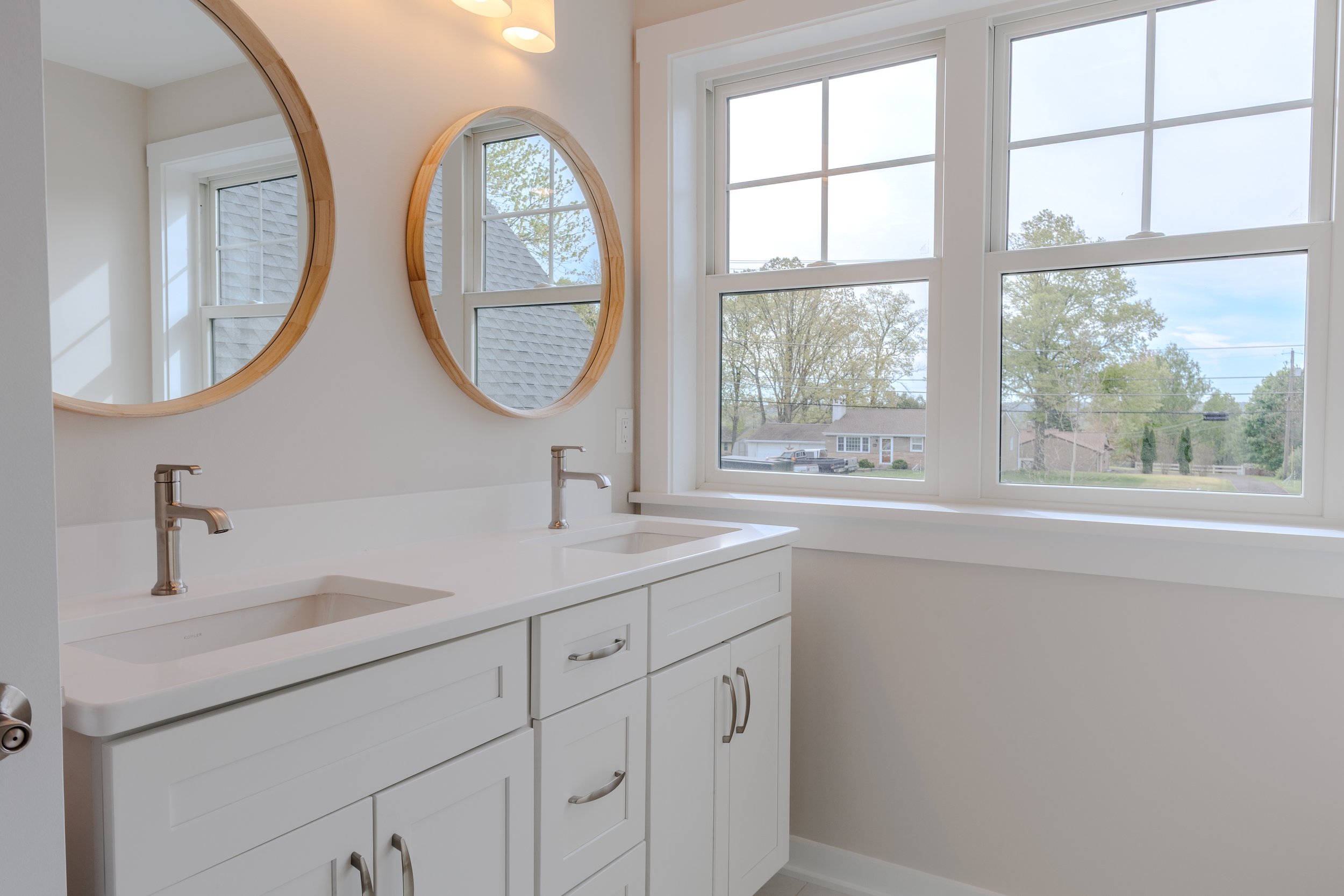 A bright bathroom with white cabinets, dual sinks with modern faucets, two round wall mirrors with wooden frames, and a large window overlooking a suburban neighborhood with trees and houses.