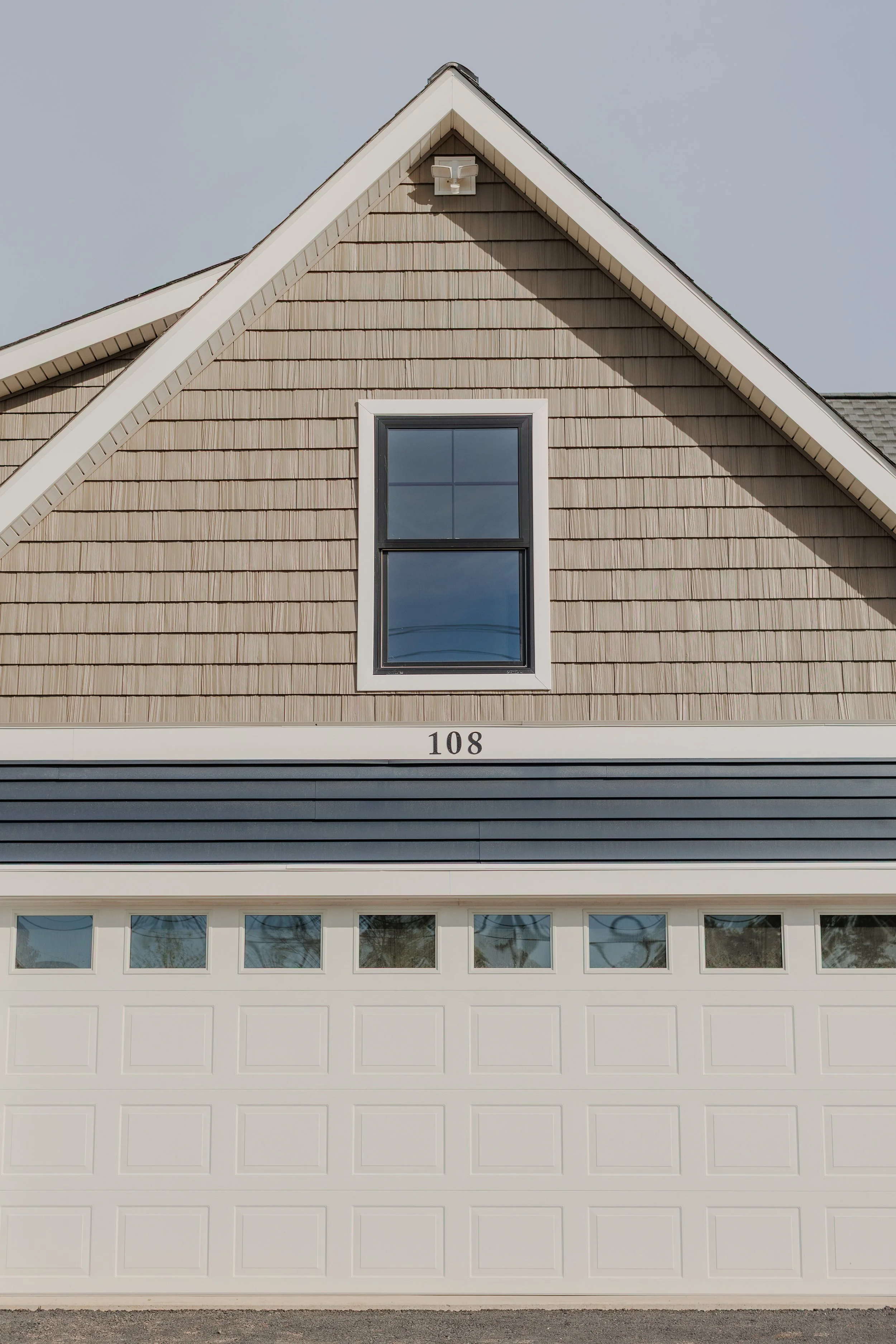 Front view of a house with beige shingles on the upper part, black window, brown garage door, and the number 108 on a horizontal white trim.