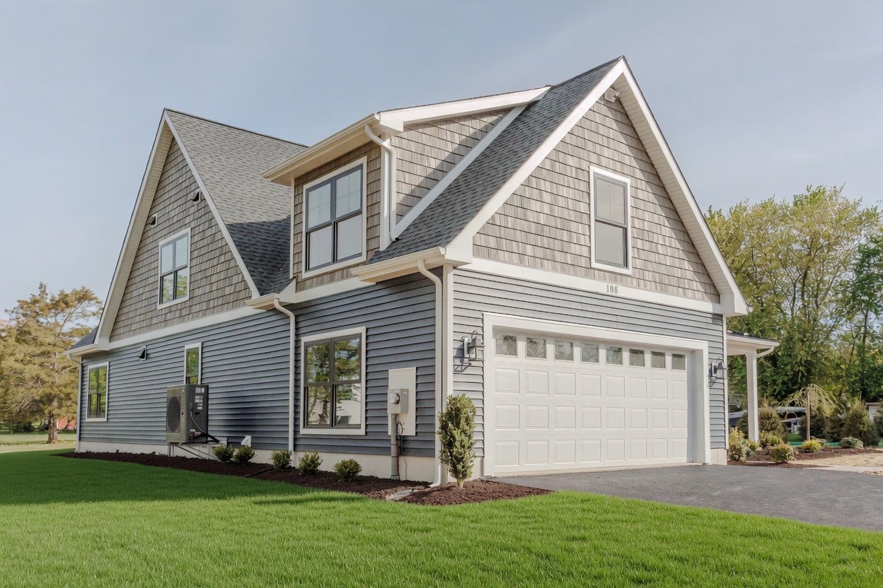 A modern two-story house with blue vinyl siding and gray shingle exterior, white trim, and a double garage. Well-maintained lawn with small bushes and trees.