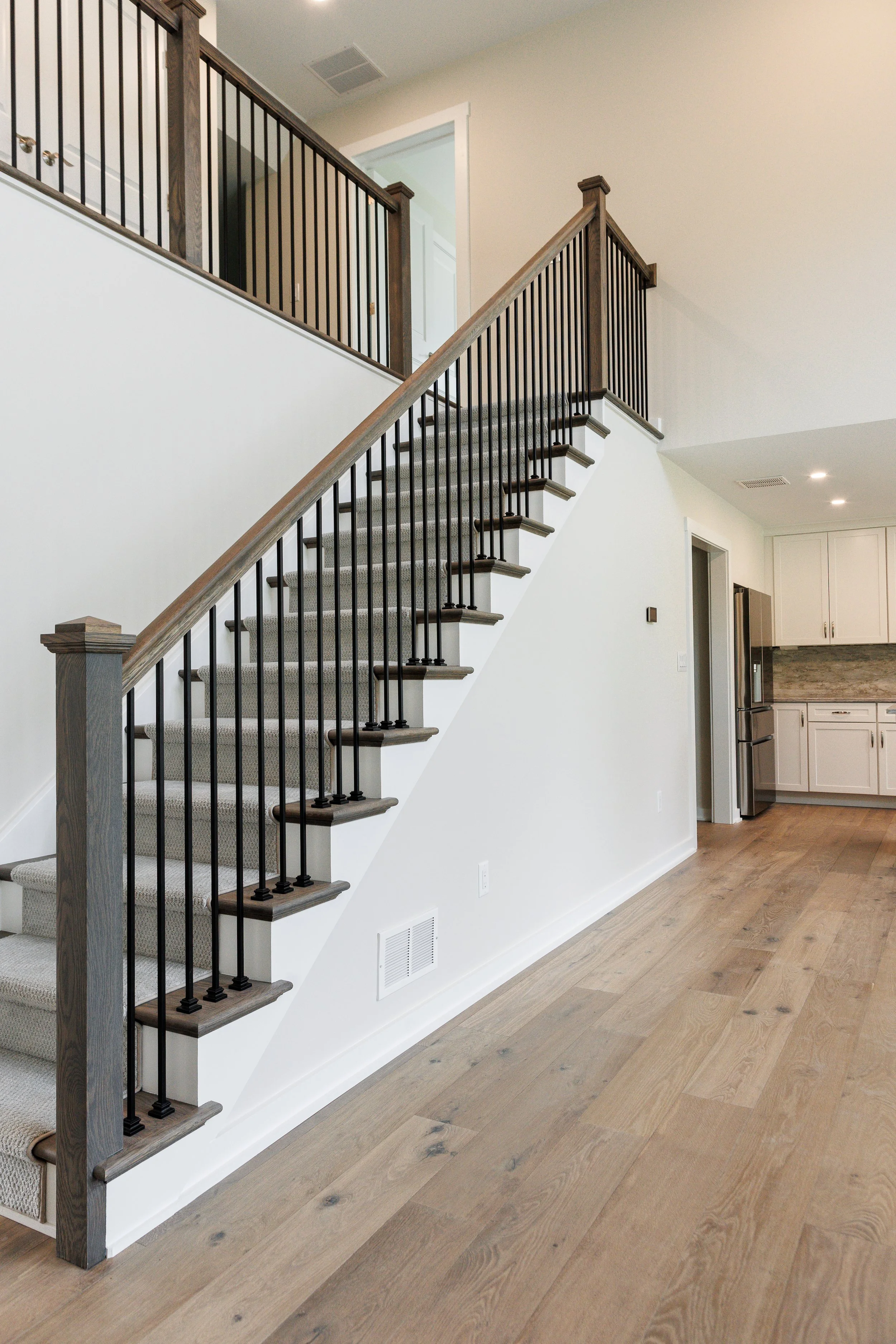 Interior view of a modern home featuring a staircase with wooden handrails and black metal balusters. Visible part of a kitchen with white cabinets and stainless steel appliances in the background, along with light hardwood flooring and white walls.