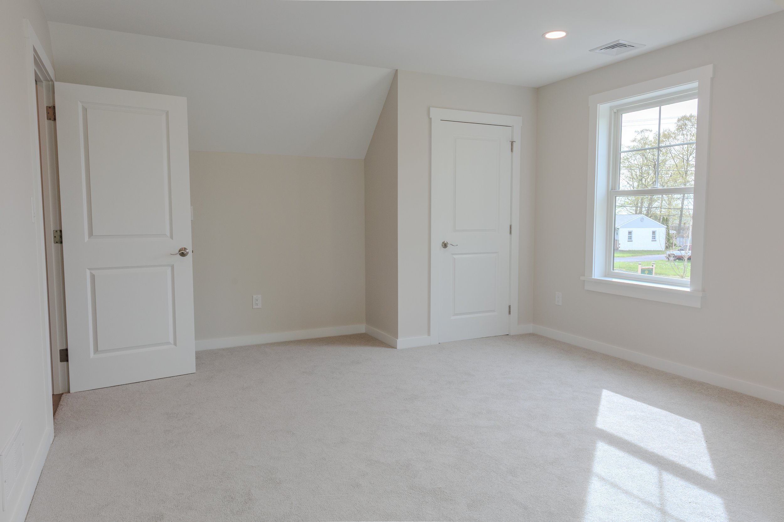 Empty room with white walls, beige carpet, two white doors, and a window showing a house and trees outside.