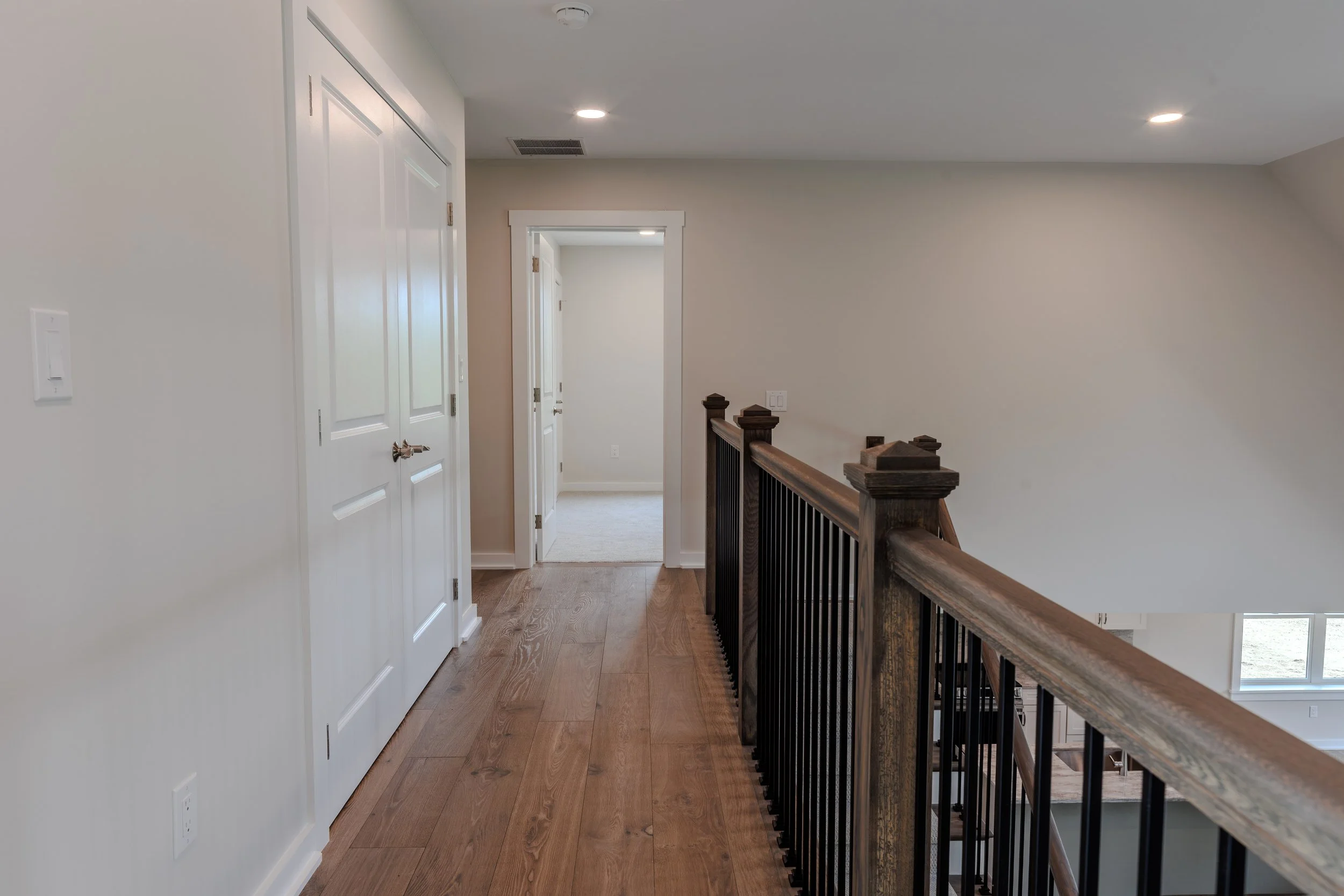 Interior view of a hallway in a house showing white doors, a wooden handrail with black balusters, beige walls, and a ceiling with recessed lighting.