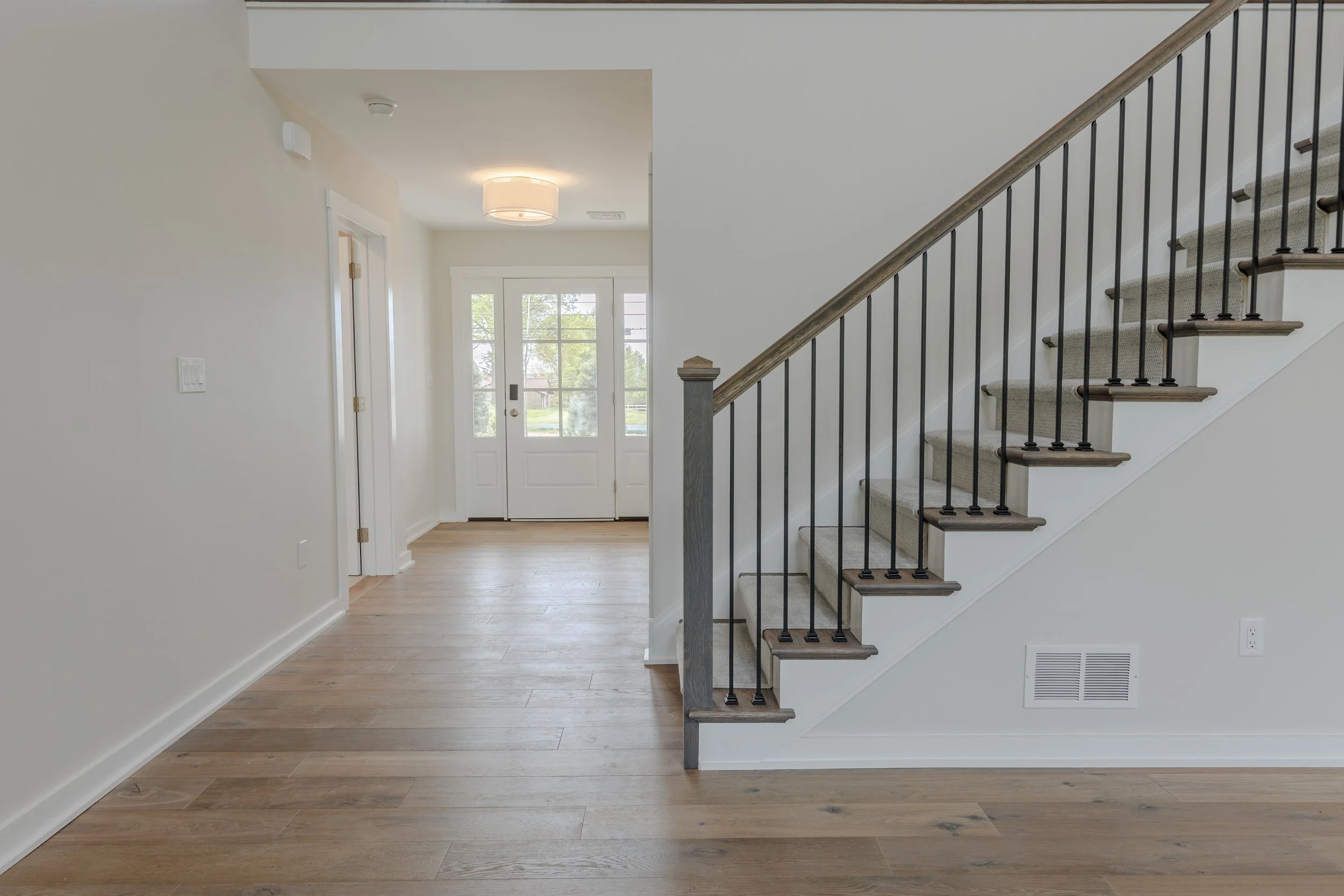Entryway of a house with a staircase on the right, white walls, wooden floors, and a front door with glass panels in the background. A ceiling light fixture is visible.