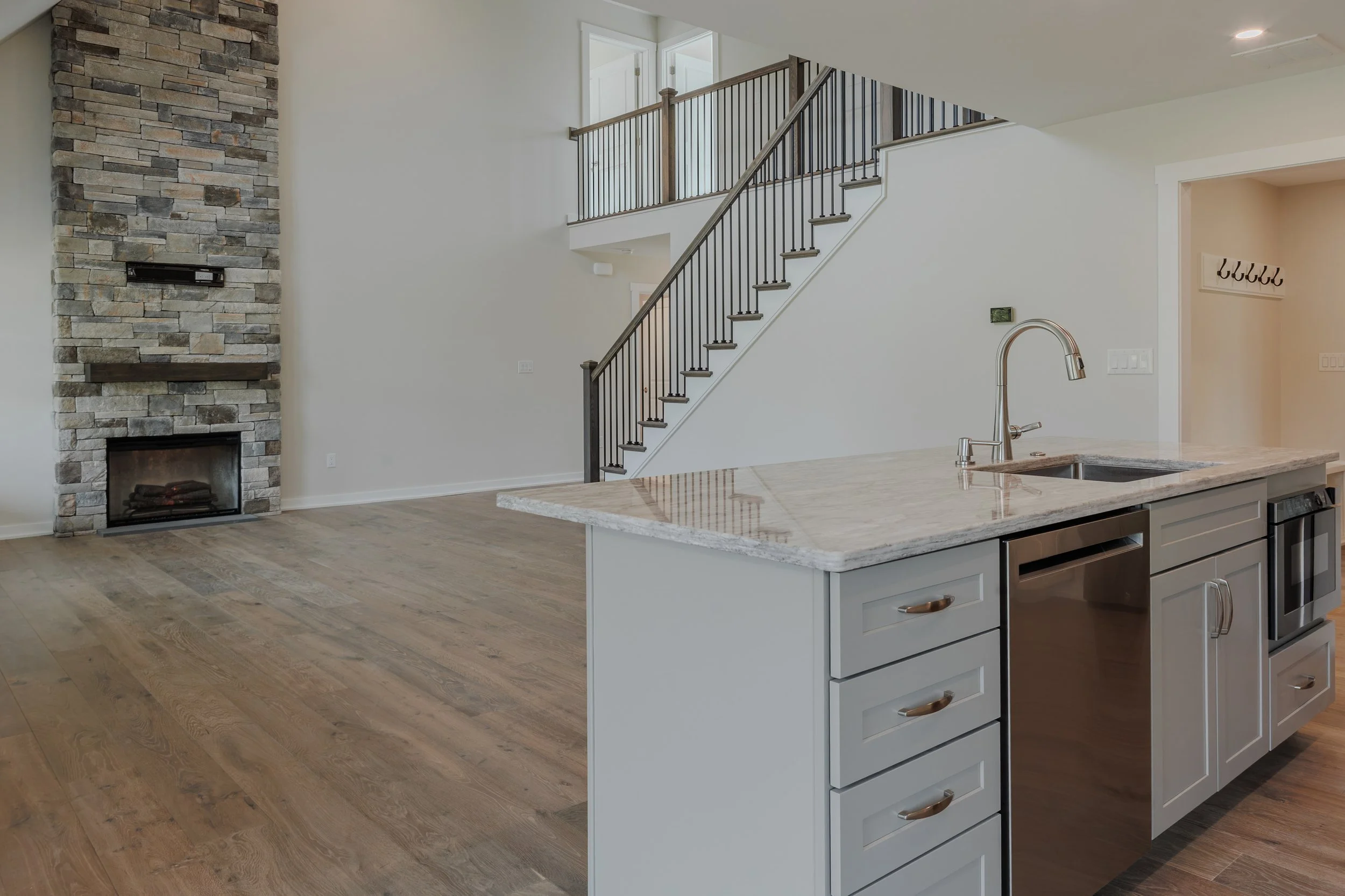 Open-concept kitchen with white cabinets, granite countertop island, stainless steel appliances, hardwood flooring, a stone fireplace, and a staircase with dark railing leading to the upper floor.