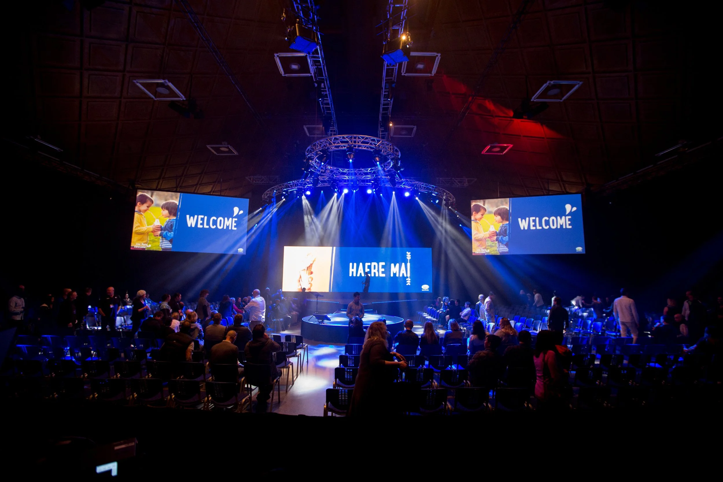 A large indoor event space with a stage illuminated by bright blue lights and large screens displaying