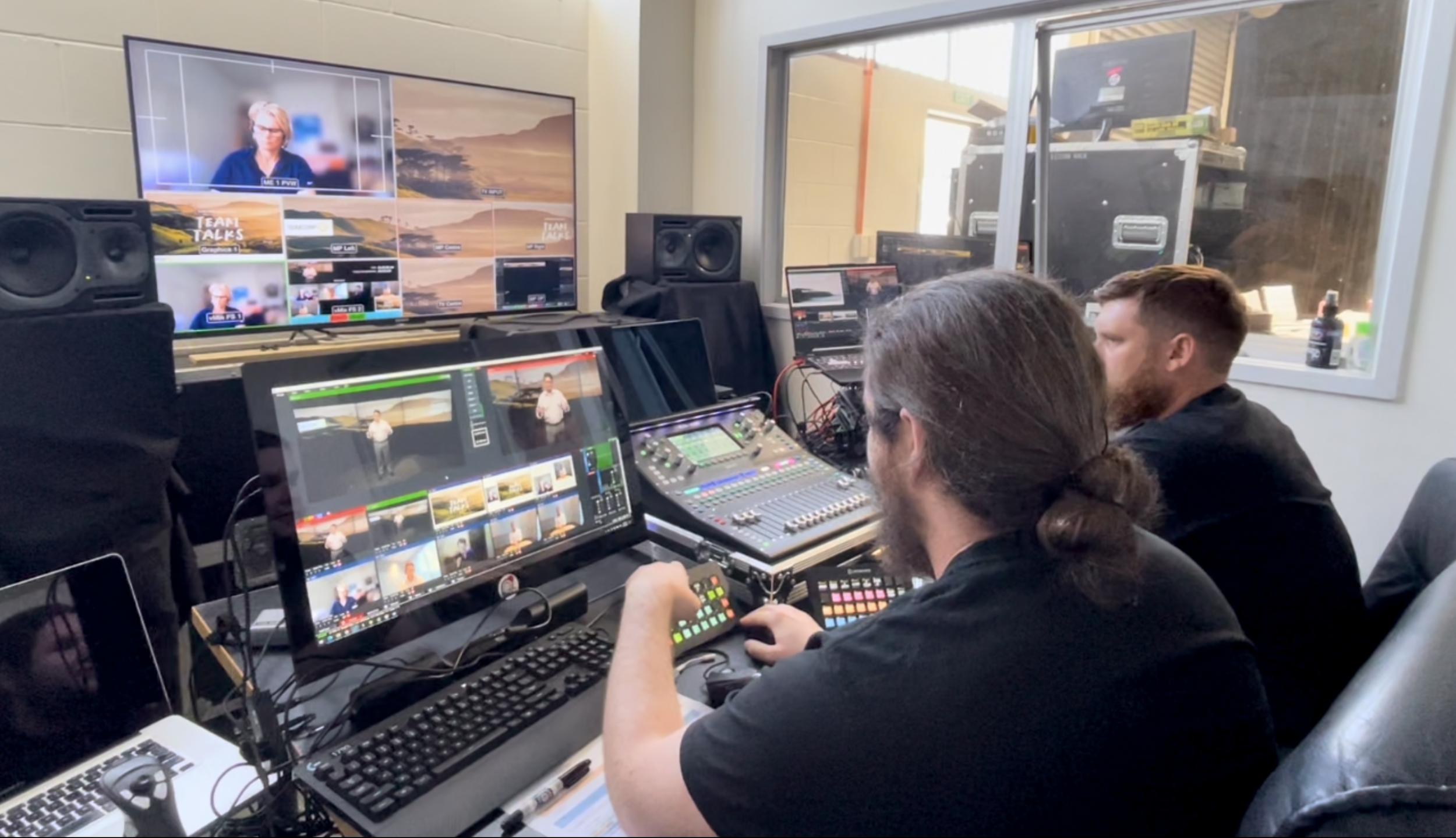 Two people in a video editing or live streaming studio, working with multiple monitors, a sound mixer, and video equipment, with a large window showing outdoor construction or storage area.