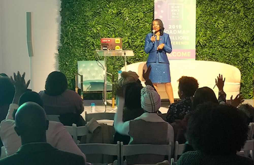 Nike exec Dr. Mona-Lisa Pinkney in blue suit on stage speaking at ‘Black Women Talk Tech’ conference, with audience raising hands, and a green living wall backdrop.