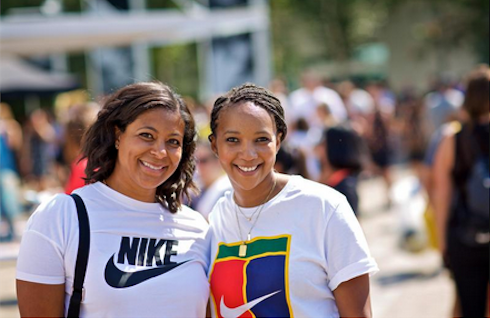 Two smiling women at an outdoor JDI Day event wearing Nike-branded t-shirts, one with classic black swoosh logo, the other with colorful geometric Nike design.