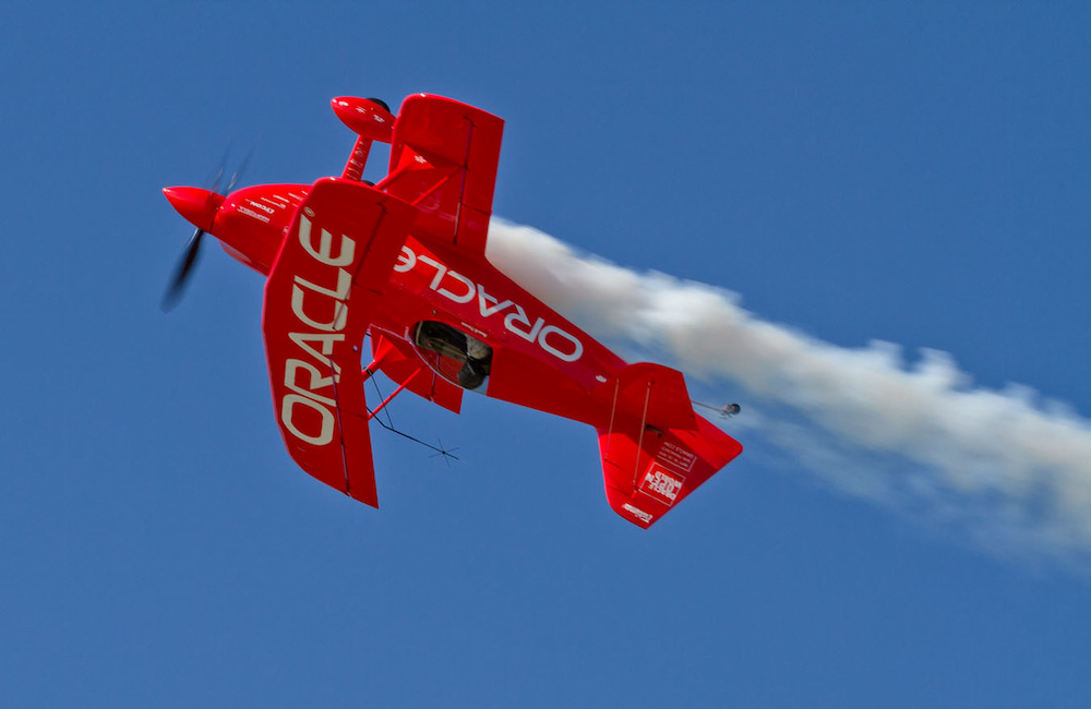 Red, Oracle-branded aerobatic plane performing barrel roll against blue sky with white smoke trail and Oracle branding visible on wings and fuselage.