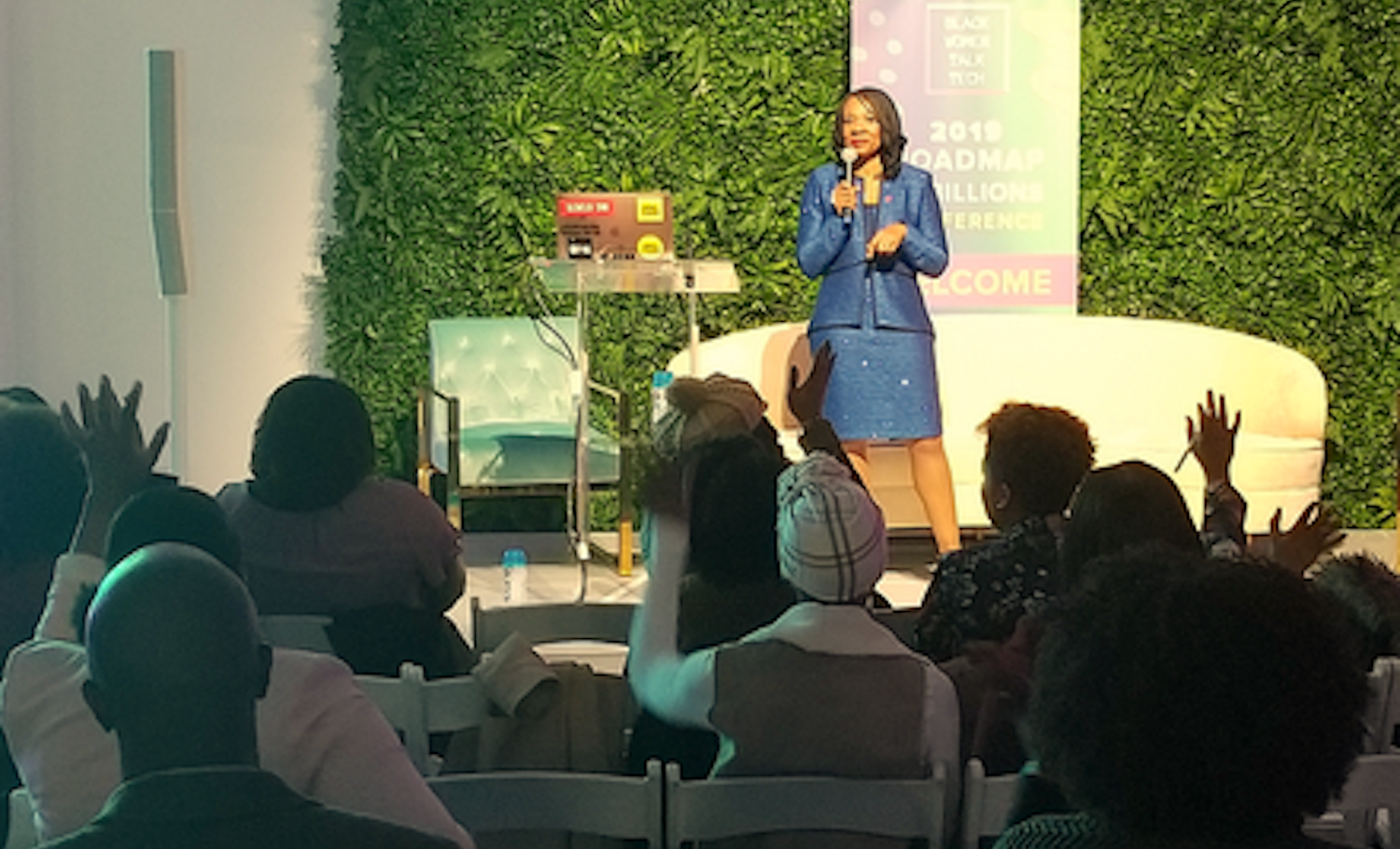 Nike exec Dr. Mona-Lisa Pinkney in blue suit on stage speaking at ‘Black Women Talk Tech’ conference, with audience raising hands, and a green living wall backdrop.