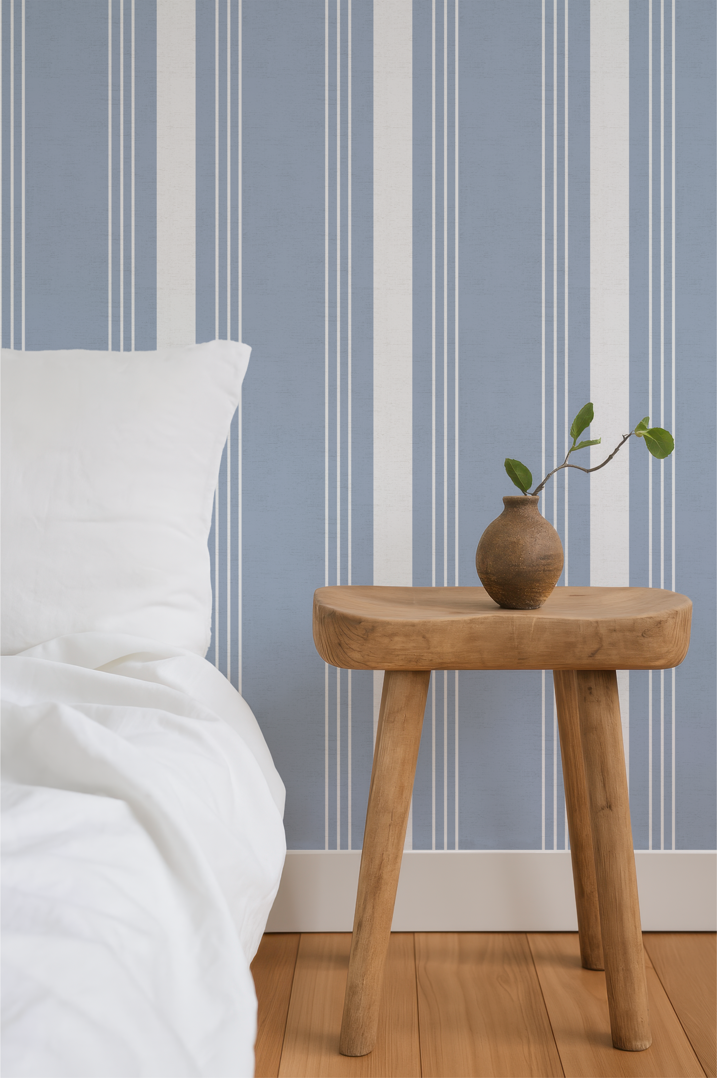 A bedroom corner with a white bed and pillow, a round wooden side table with a small brown ceramic vase containing a branch with green leaves, a blue and white striped wall, and hardwood flooring.