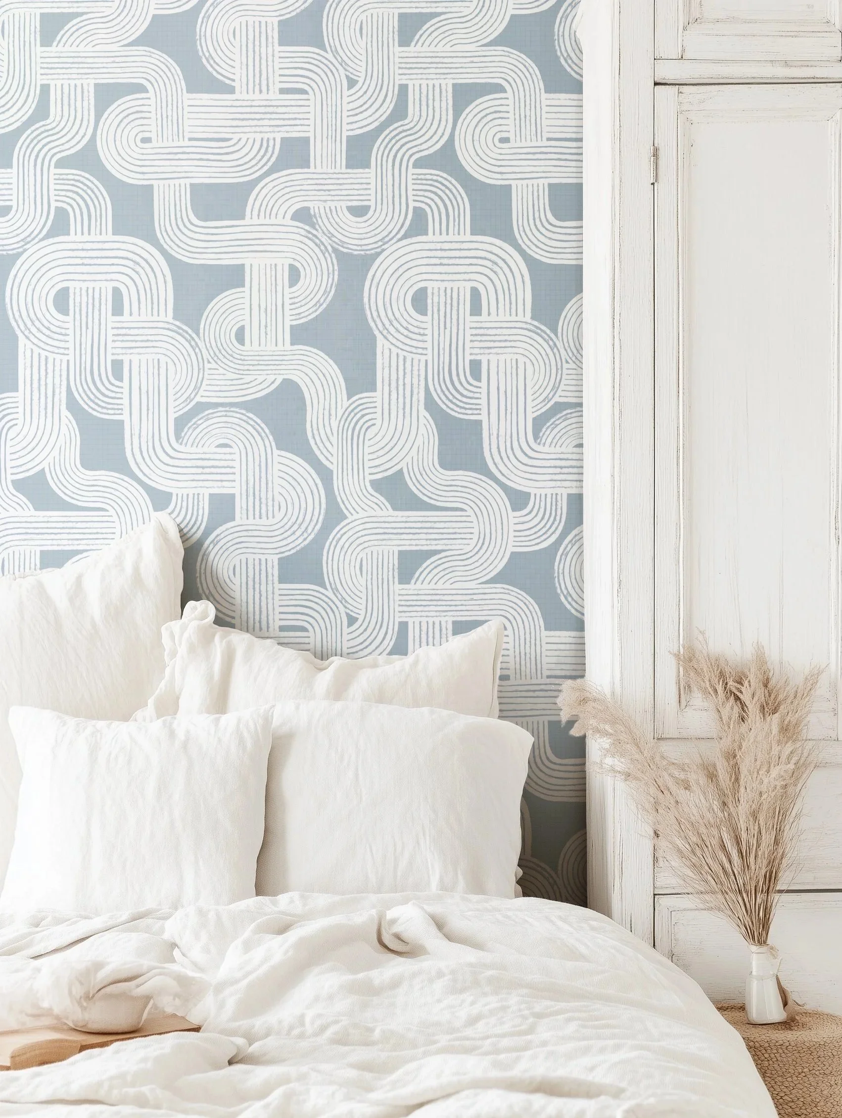 A cozy bedroom with a white bed, soft white linens, a distressed white wooden headboard, bold geometric wallpaper in light blue and white, and a neutral-toned fuzzy rug with a tall dried pampas grass arrangement in a white vase.