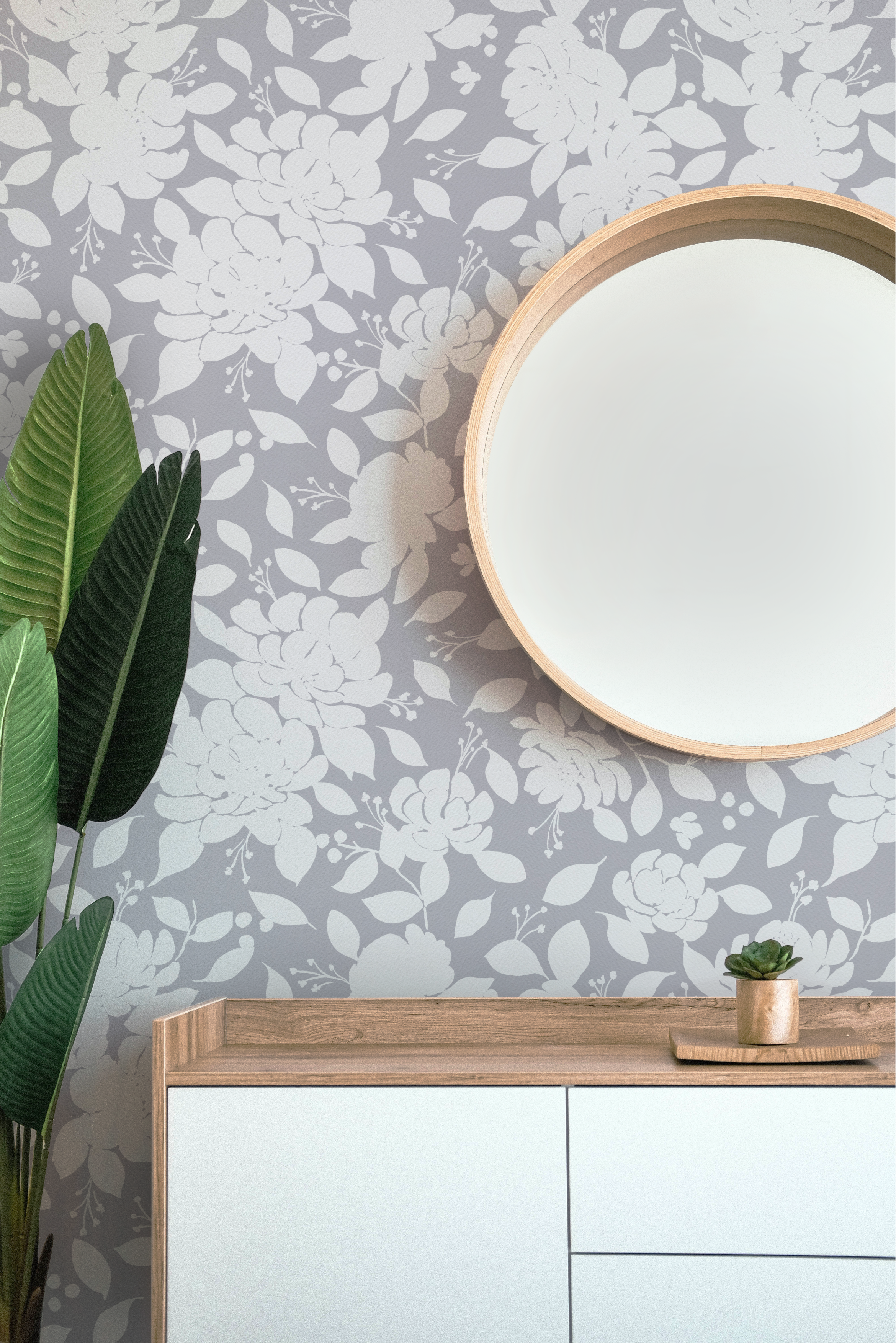 A decorative wall with gray and white floral wallpaper, a round mirror with a wooden frame, a wooden table with a white front, and a small potted succulent plant in a wooden container on the table. There are green leaves from a large plant on the lef