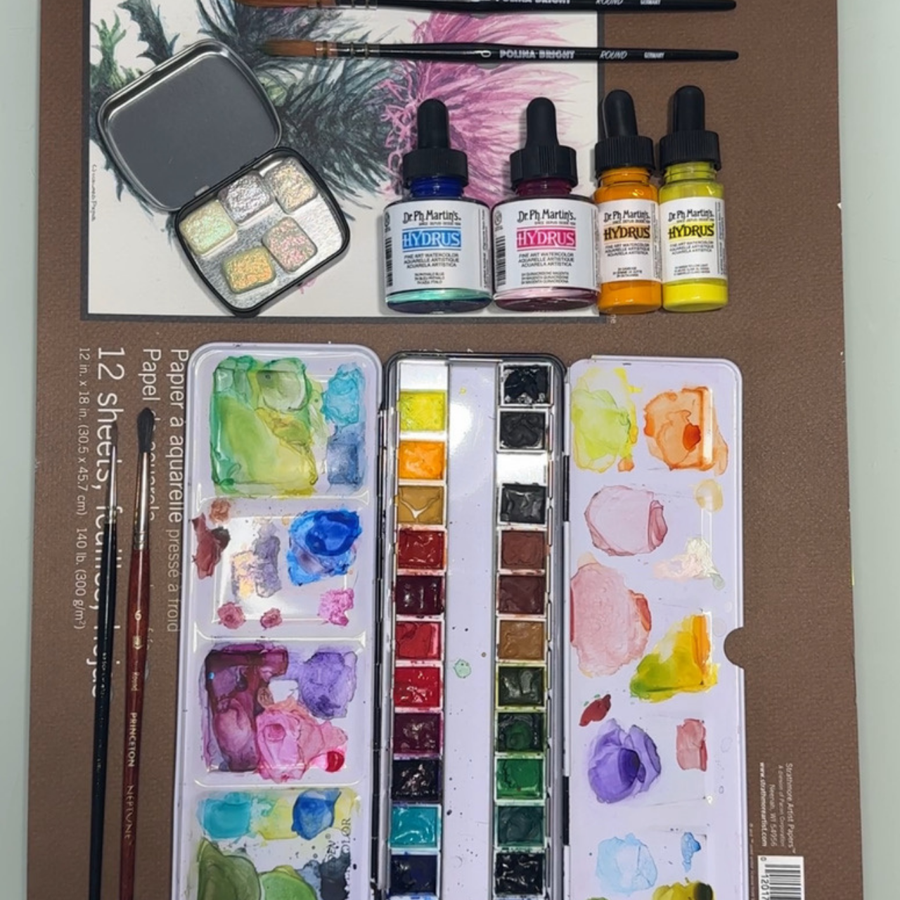 Watercolor painting supplies, including a palette with various colors, three bottles of Dr. Ph. Martens' Hydrus watercolor paint in blue, pink, yellow, two paintbrushes, and a paper watercolor set with colorful paint splashes on a brown and white paper mat.