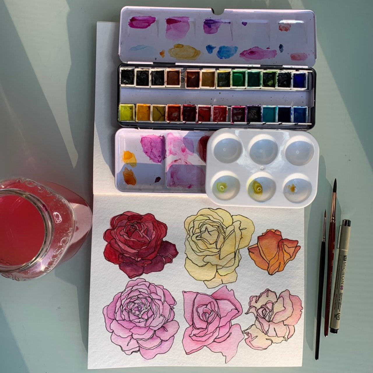 Watercolor paints, brushes, and sketches of roses in various colors on paper, with a glass of water on the side.