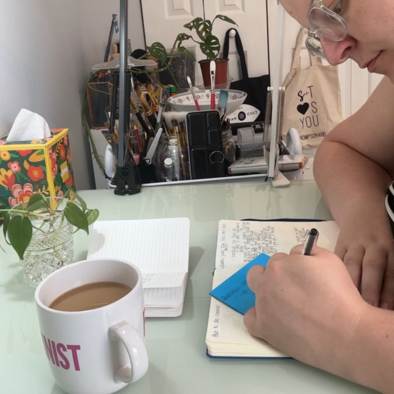 A person is sitting at a glass table writing in a notebook with a pen. On the table, there is a cup of coffee, a small notepad, and a potted plant. In the background, a makeup or art station with brushes, tools, and a plant in a small pot is visible, along with a tote bag hanging on the wall that says "S+T ♥ YOU." The person is wearing glasses and has light skin.