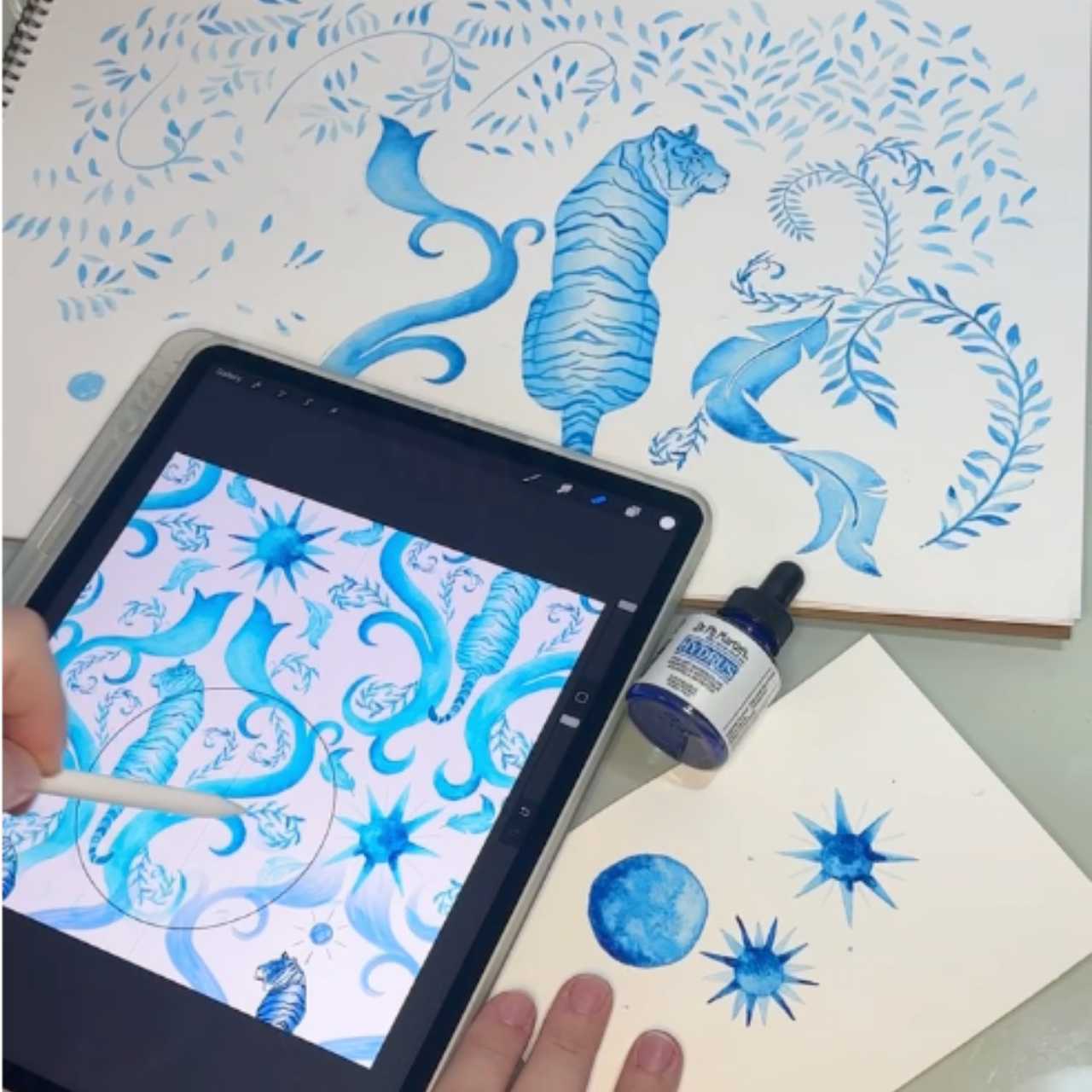 A person is creating a blue ink illustration of a tiger in the jungle with swirling foliage and stars on paper, using a digital tablet and stylus. An ink bottle is nearby.