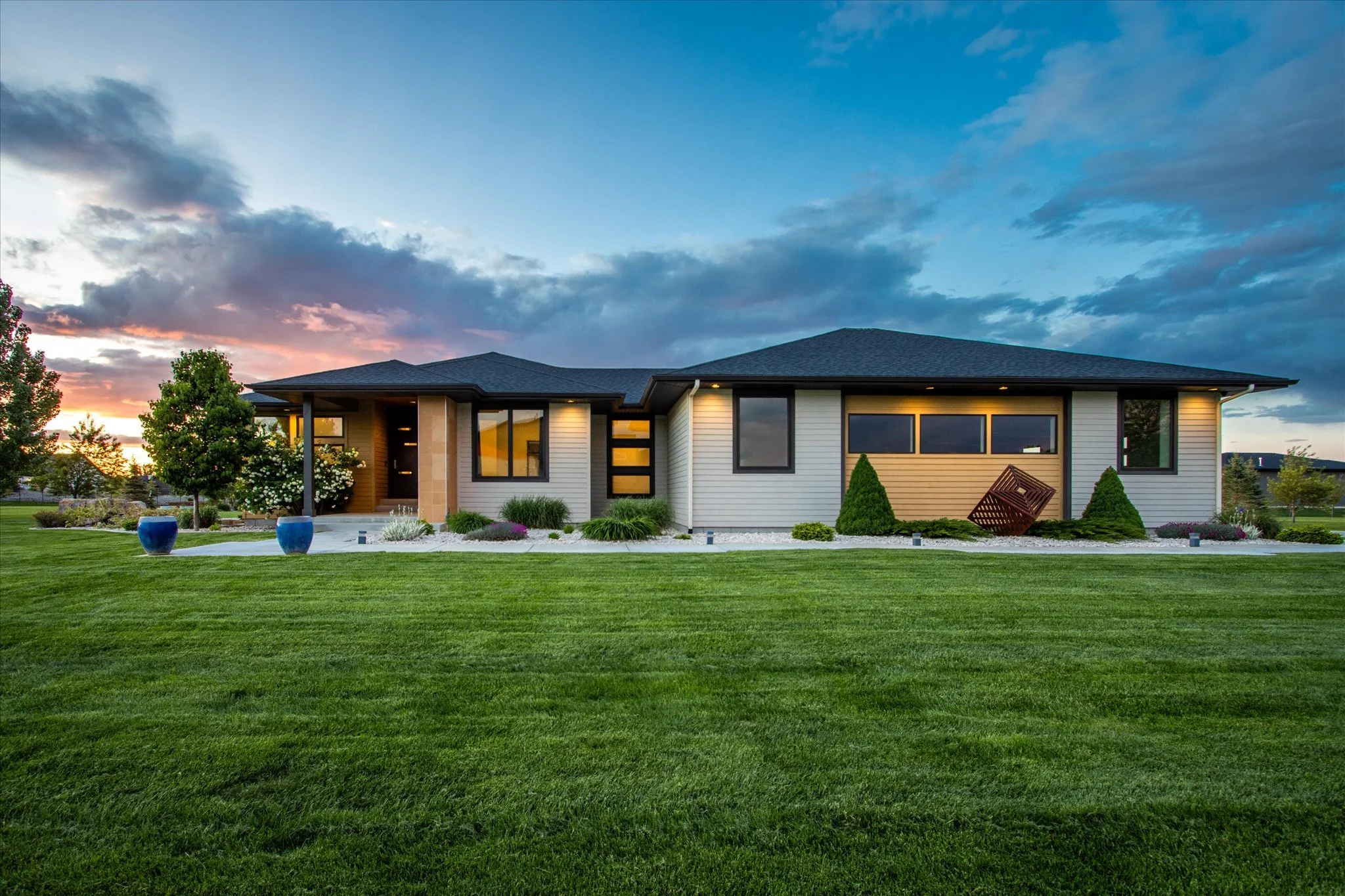 Modern house with large lawn and landscaped garden at sunset, featuring multiple large windows, shrubbery, and decorative planters.