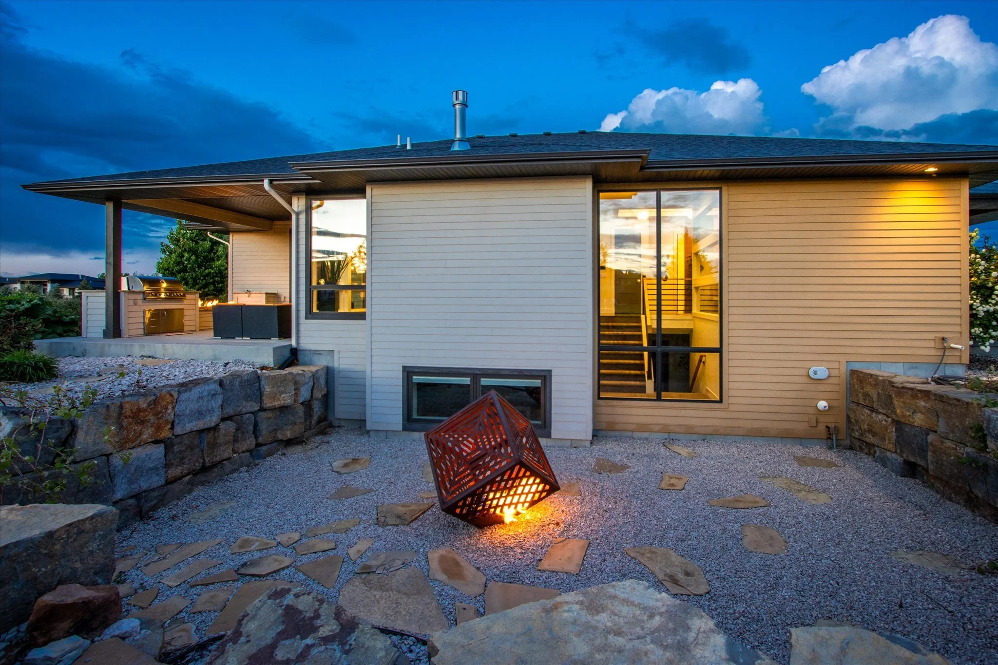 Backyard of a modern house with a stone patio, outdoor fireplace, and large glass door leading inside, during sunset.
