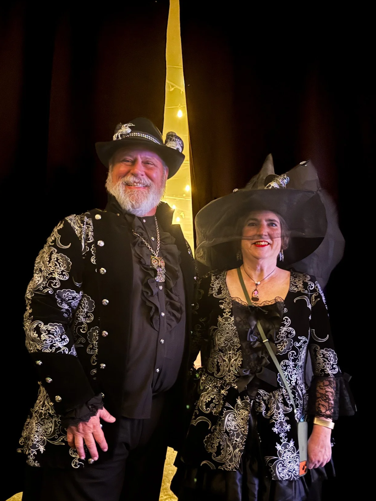 A man and woman dressed in elaborate black and white costumes with witch hats, posing together at a festive event.