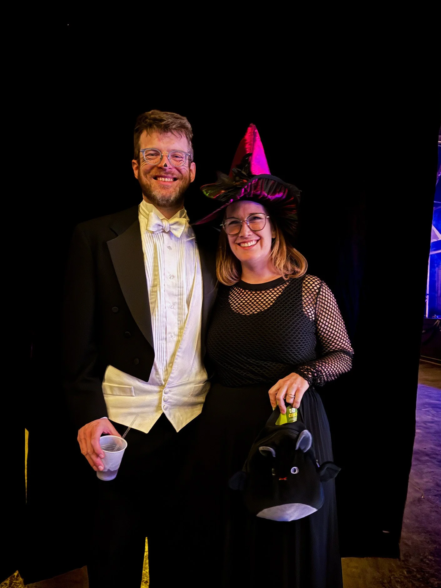 A man and woman dressed in festive costumes, smiling at the camera. The man is in a tuxedo with a white bow tie, holding a cup, and the woman is wearing a large pink and black witch hat and a black fishnet top.