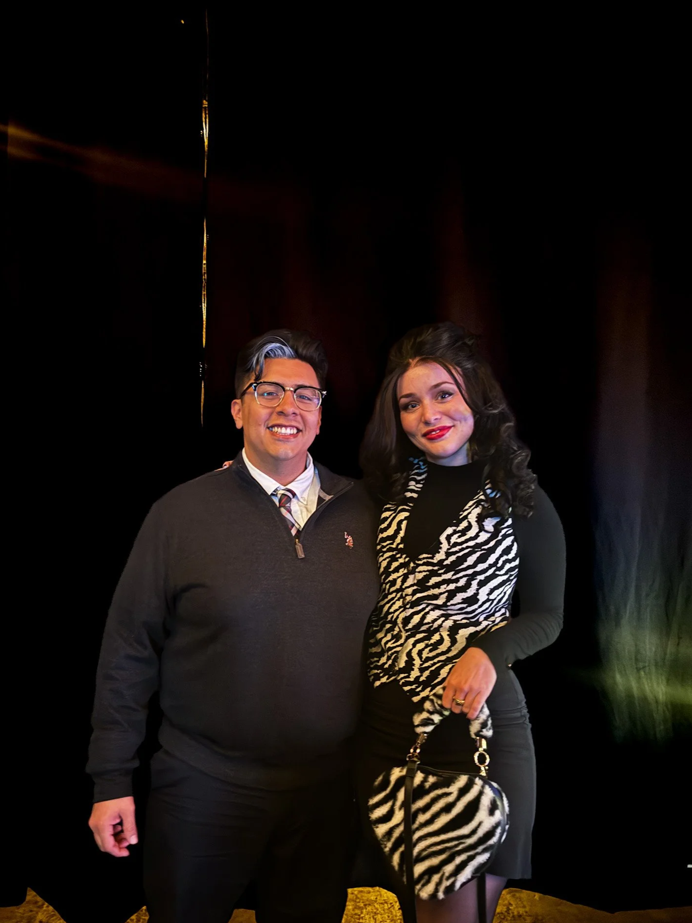 A man and woman standing together in front of a dark background, smiling at the camera. The man is wearing glasses, a dark sweater, and a collared shirt with a tie. The woman has long dark hair, wearing a black top and a zebra print scarf, and is hol